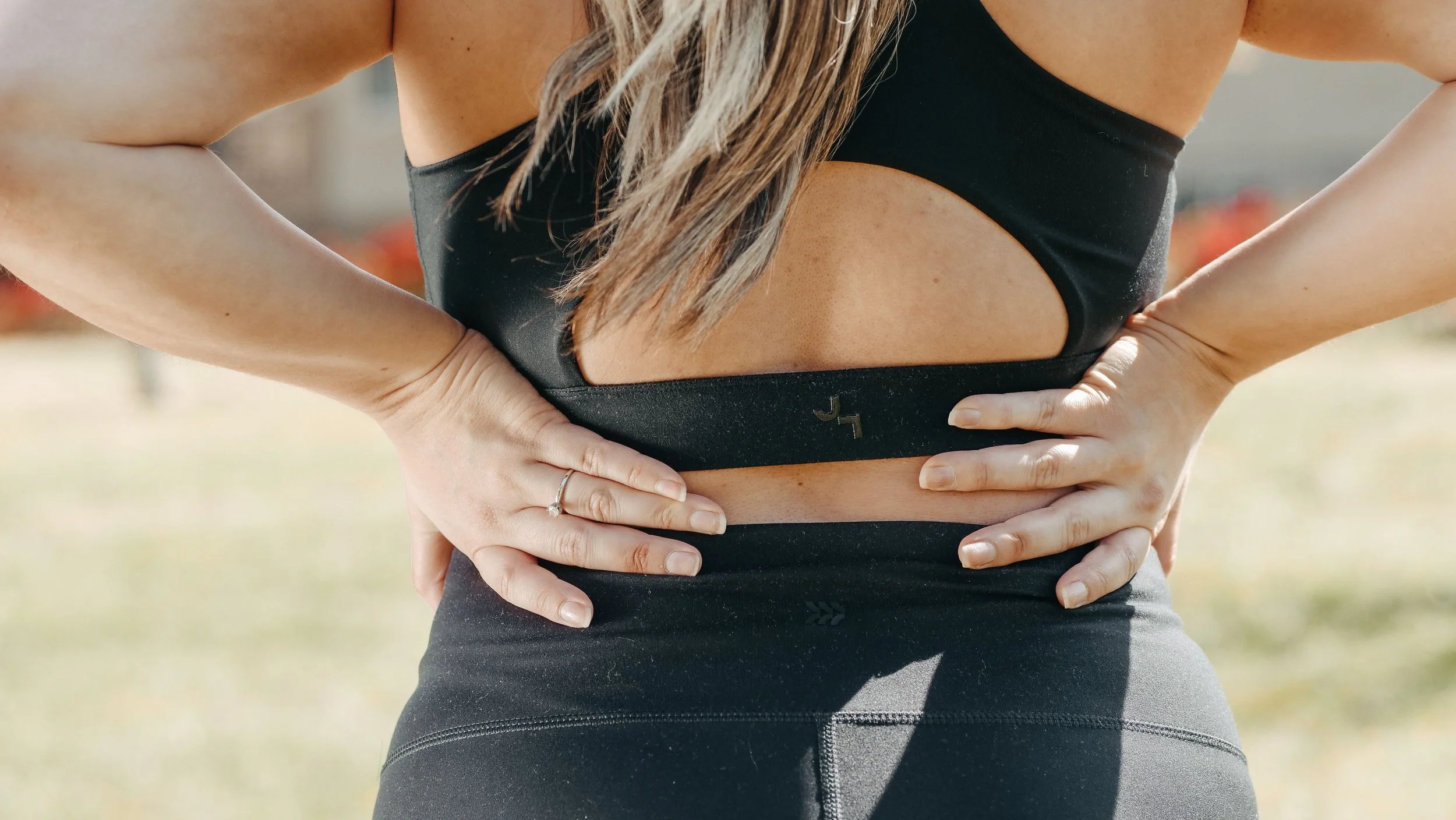 Person in black athletic wear with hands on hips outdoors, with long hair and a ring on their left hand.