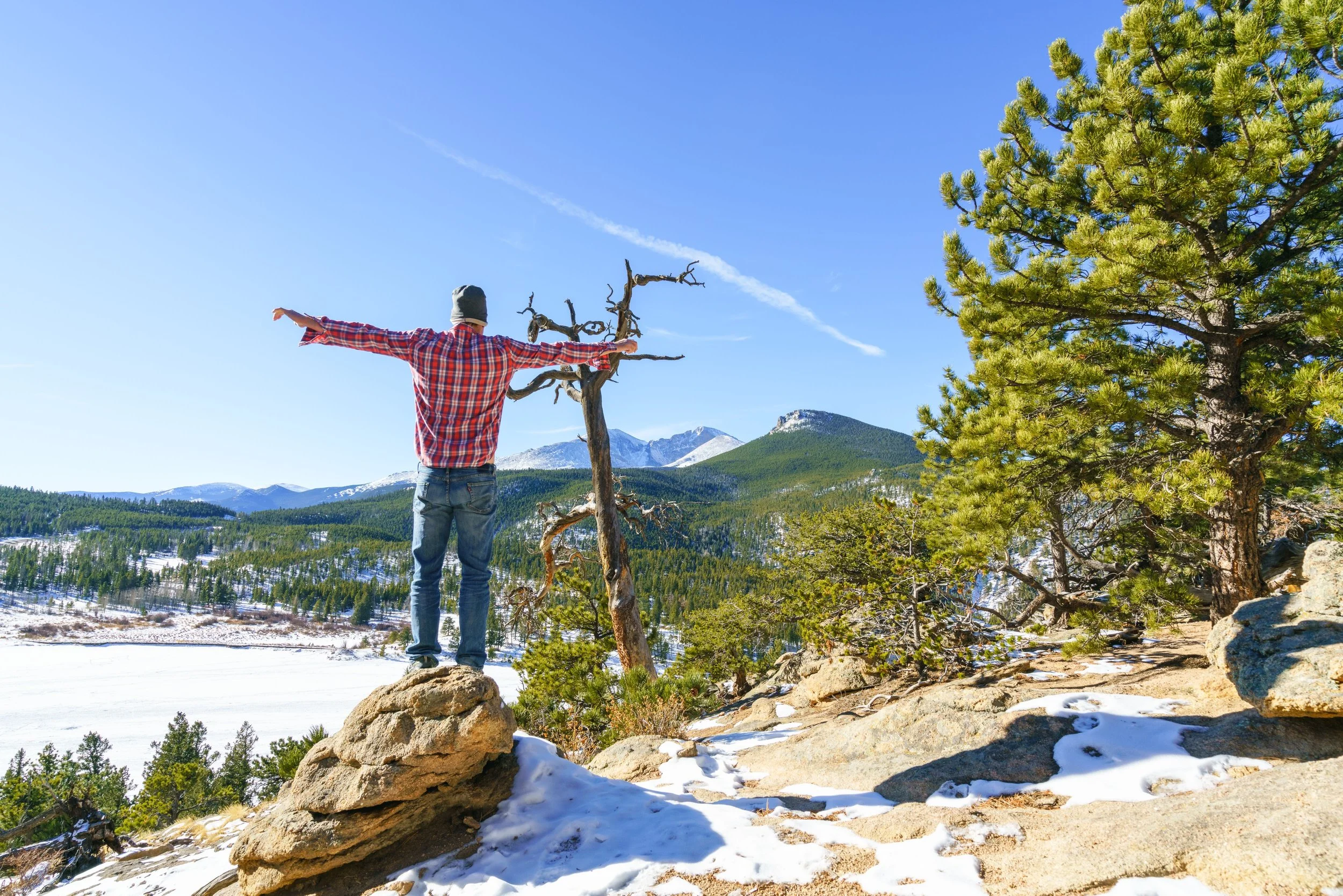 Person in a red plaid shirt and gray beanie standing on rocks with arms outstretched, facing a scenic mountain landscape with snowy peaks, evergreen trees, and a bright blue sky.