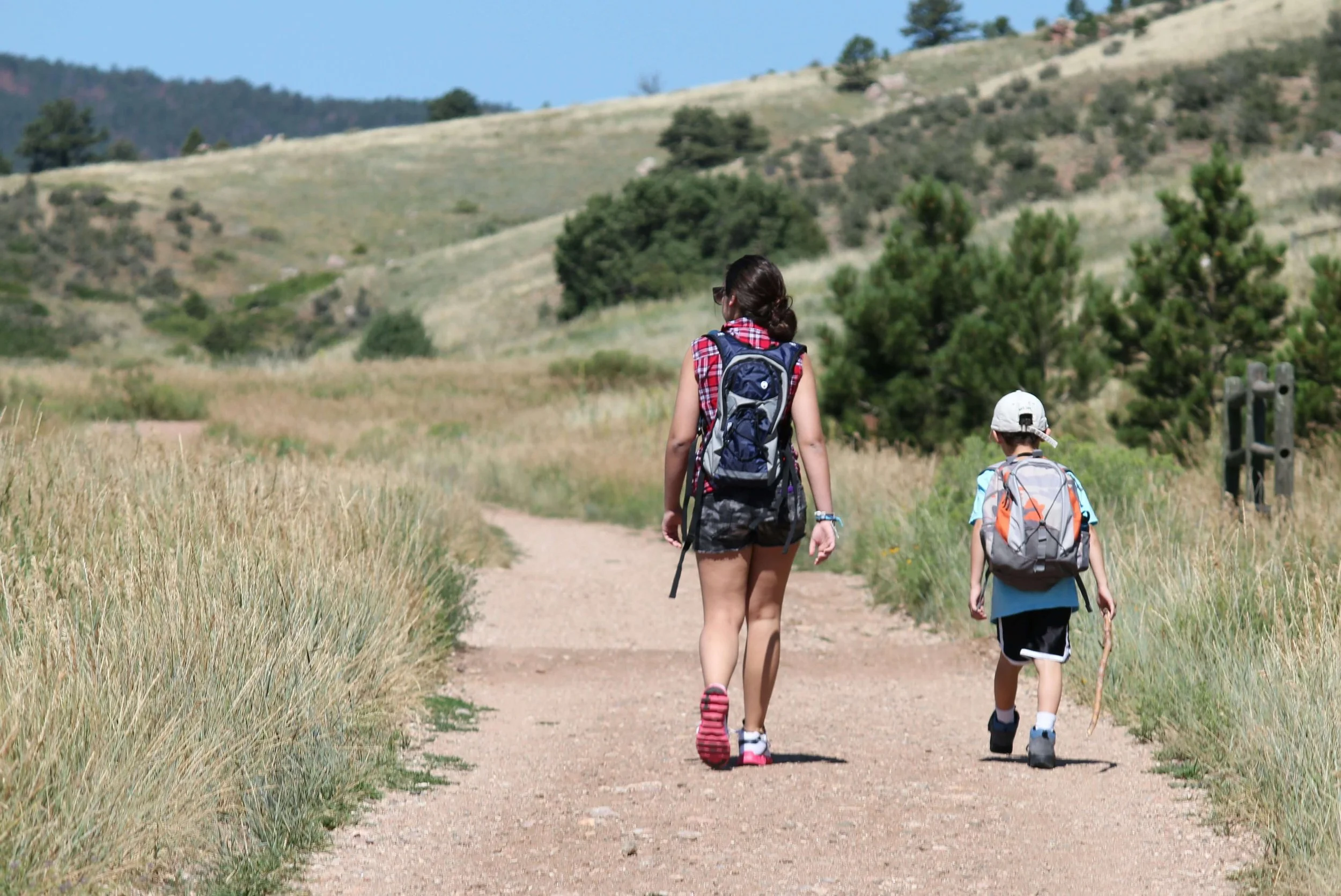 A woman and a young boy walking on a dirt trail through a grassy field with hills and trees in the background on a sunny day.