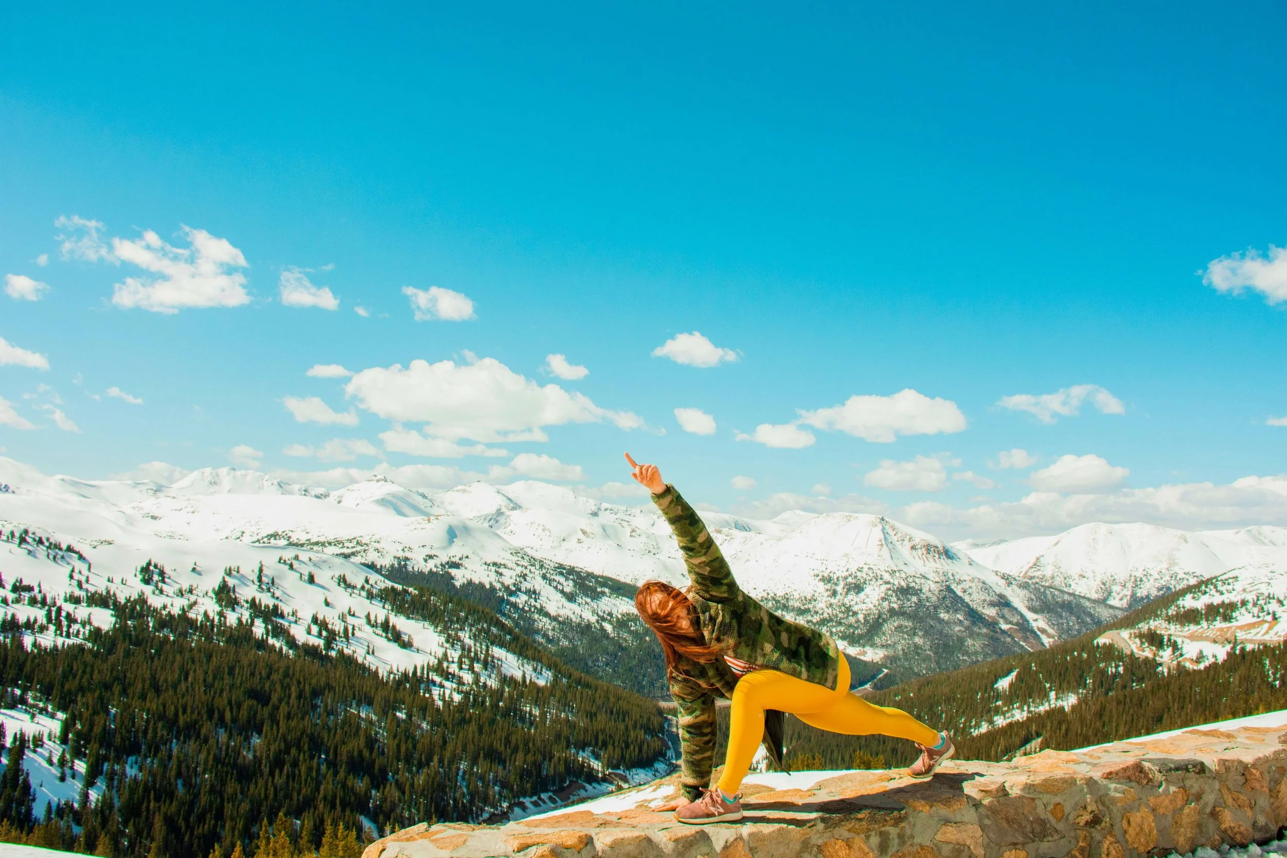 Person in colorful clothing performing a yoga pose on a stone ledge in snowy mountainous landscape with forests and a blue sky with clouds.