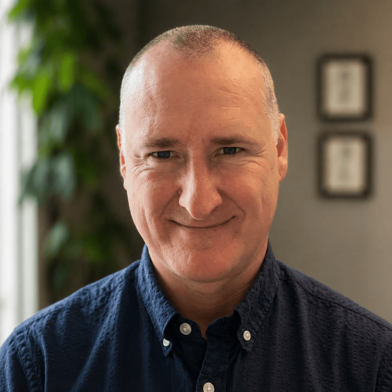 Close-up portrait of a middle-aged man with a shaved head, smiling slightly, wearing a dark blue button-up shirt, in an indoor setting with a blurred background of framed pictures and a leafy plant.