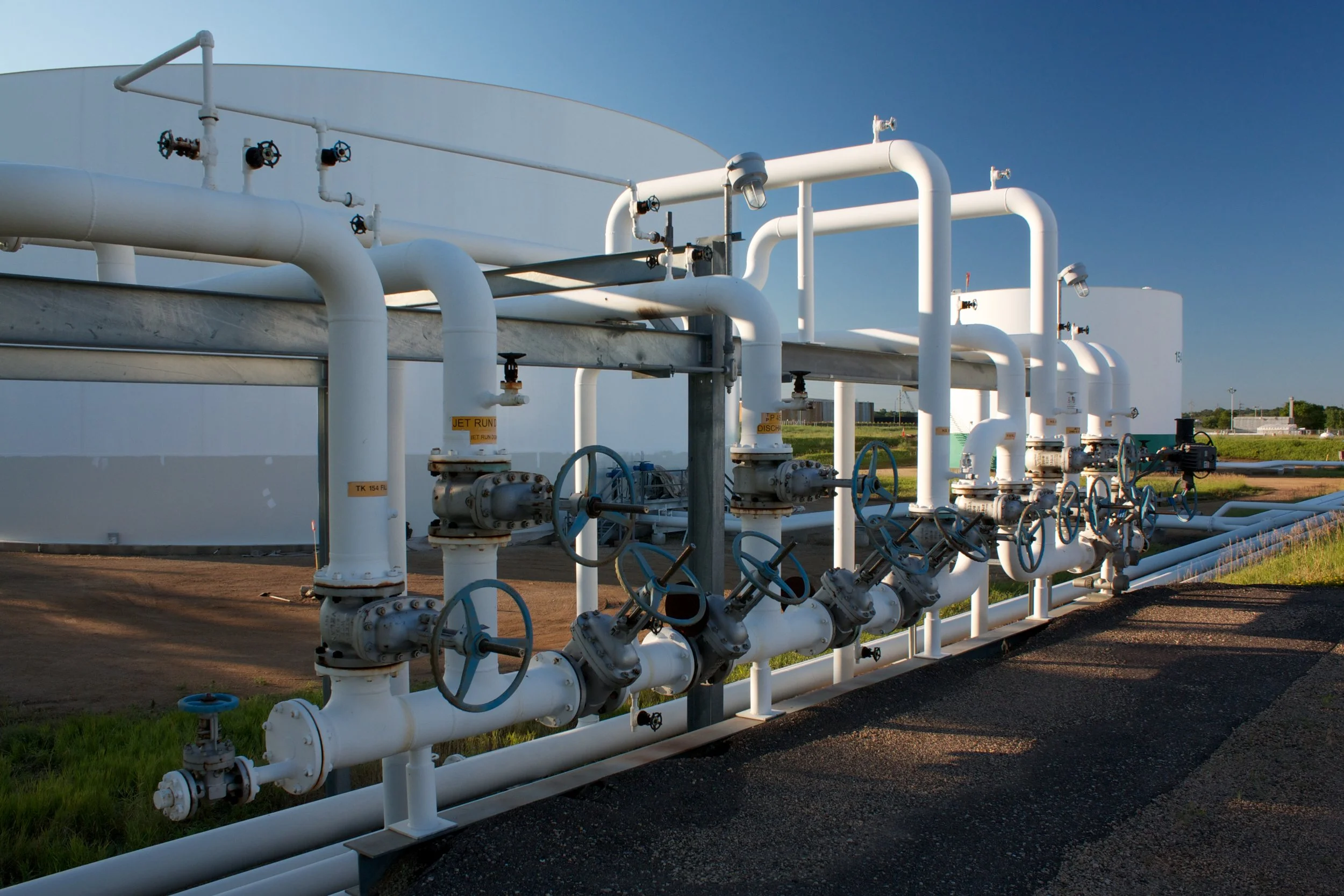 Industrial piping system with large white pipes and blue valves outdoors near storage tanks.
