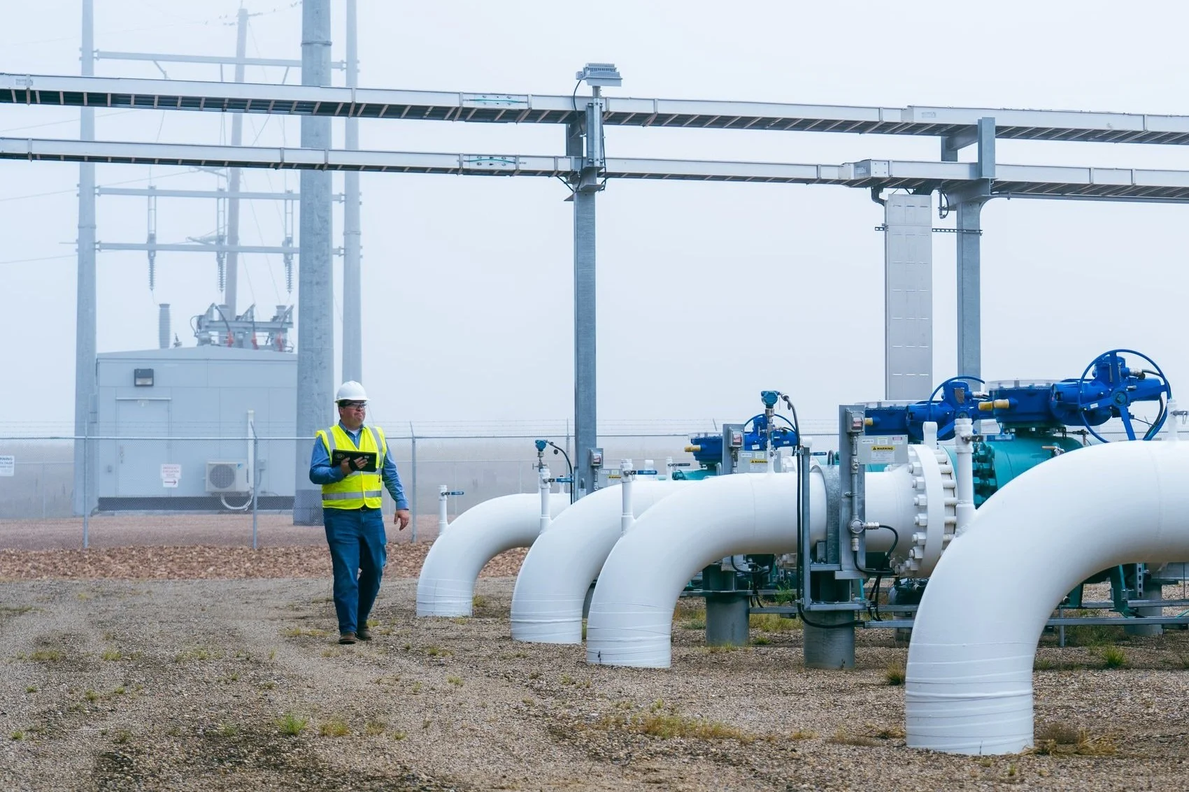 Industrial facility with a worker in safety gear walking near large white pipes and blue valves.