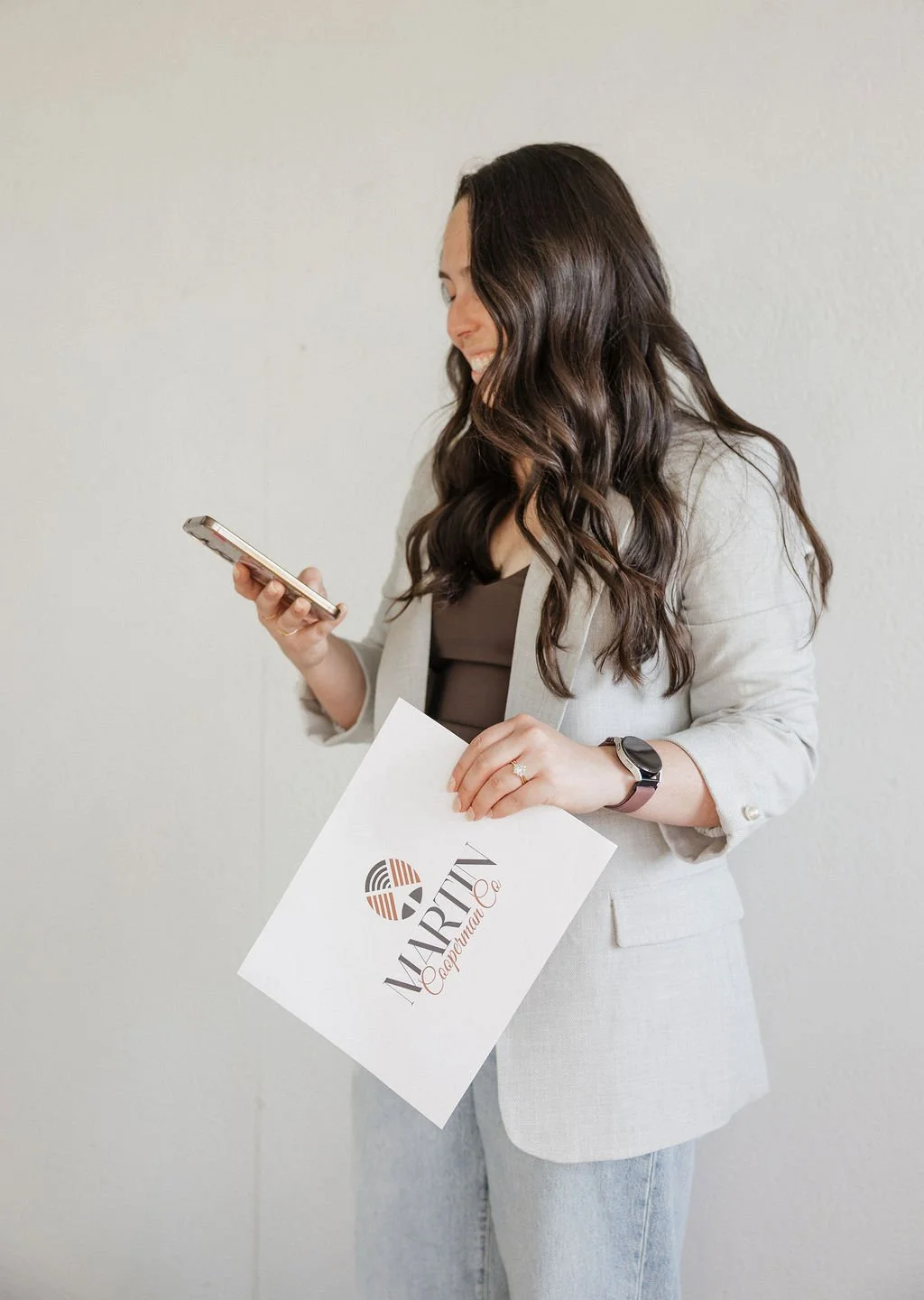 A woman with long, wavy brown hair smiling and looking at her smartphone, holding a business card and a paper with a logo that says 'MARIT Capital'.