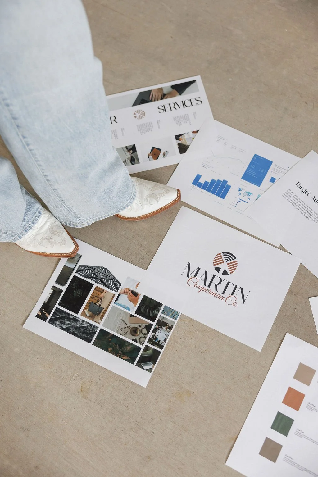 Various printed marketing materials and photographs scattered on the floor, with a person's legs visible, wearing jeans and cowboy boots.