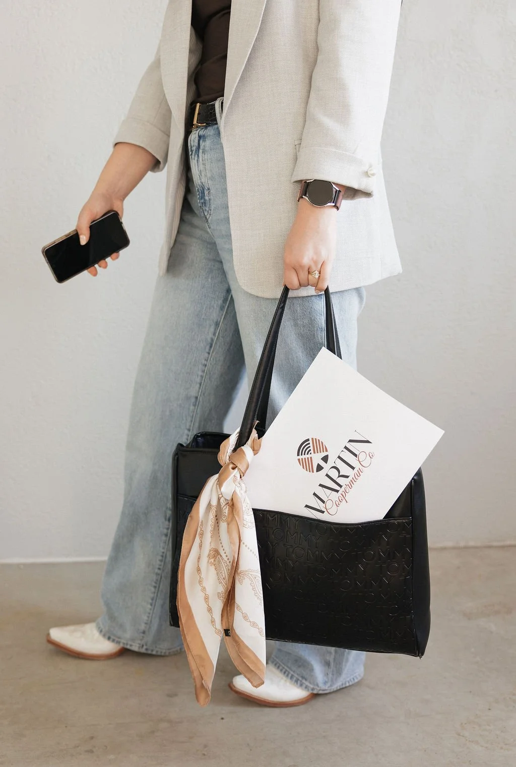 Person carrying a black tote bag with a white card and a beige scarf tied to the handle, holding a smartphone, wearing a beige blazer, light gray jeans, white shoes, and a smartwatch.