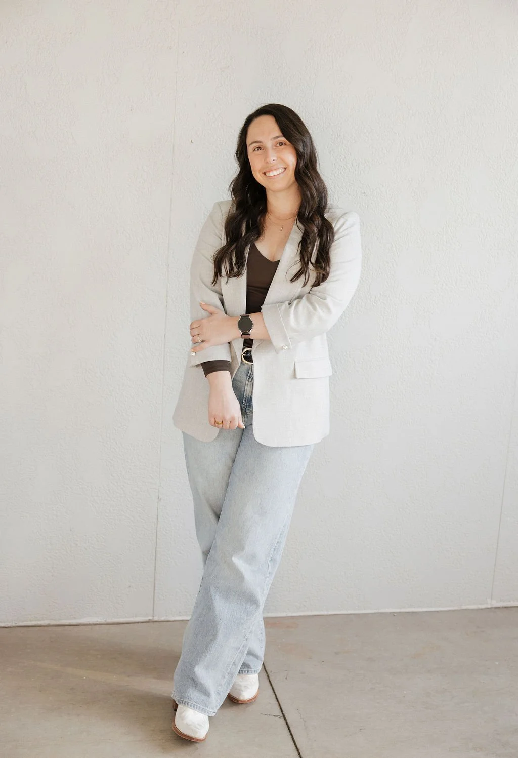 Woman with long dark wavy hair wearing a light-colored blazer, black top, light wash jeans, white shoes, and a watch, smiling with arms crossed, standing against a plain light-colored wall.