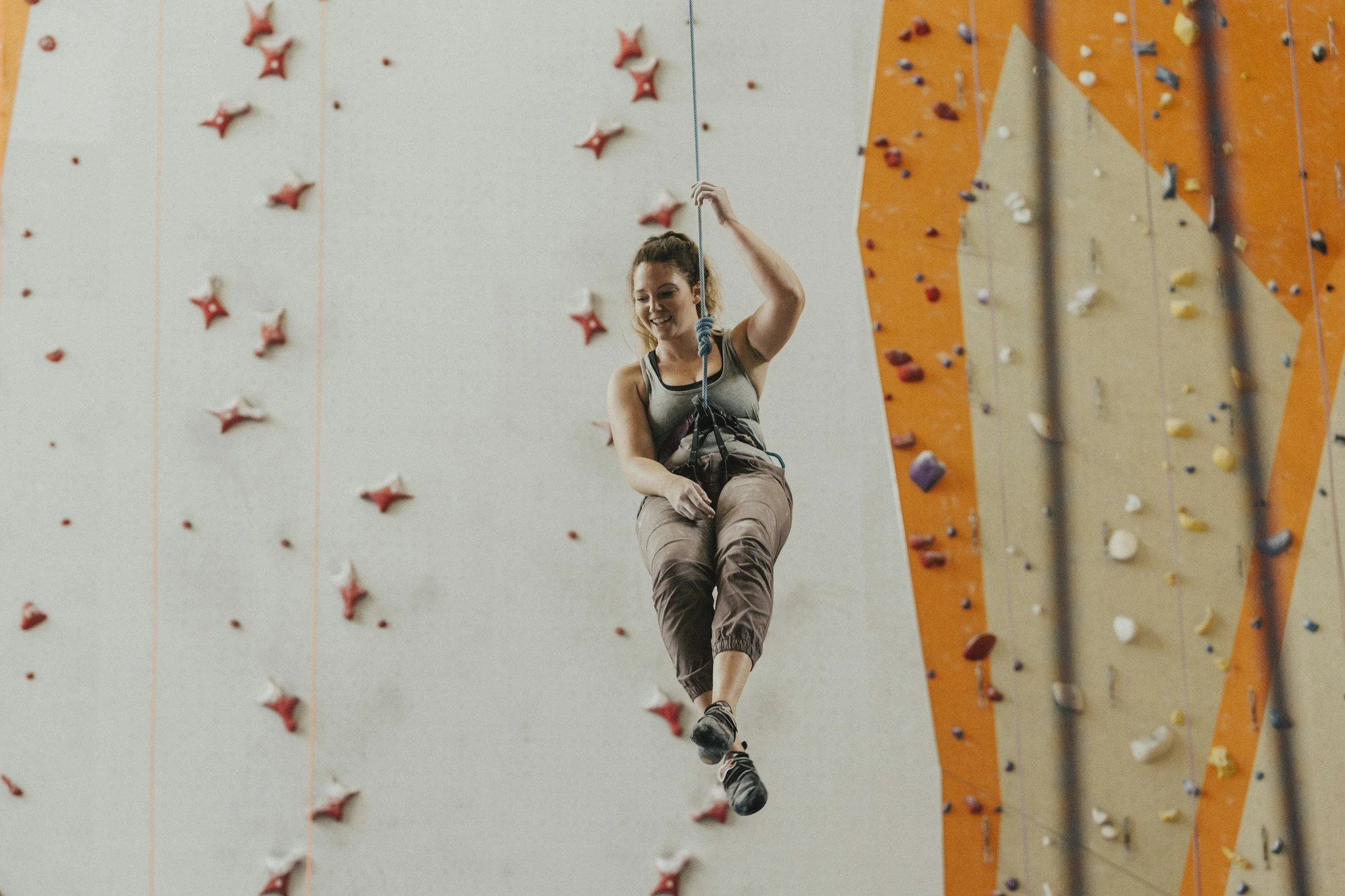 Woman on a harness and rope descending in an indoor rock climbing gym with climbing walls and holds visible.