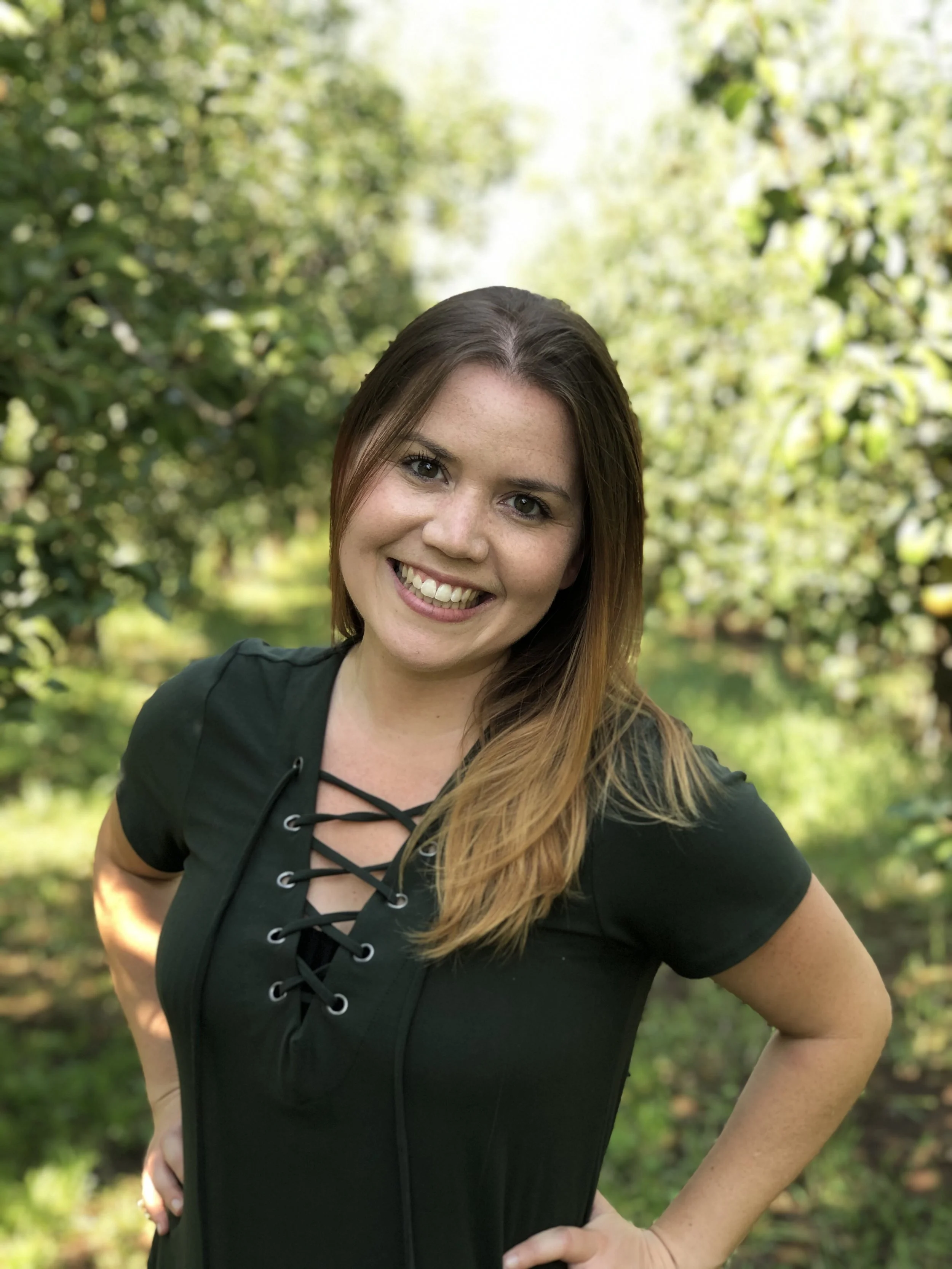 A smiling woman with long brown hair standing outdoors in a lush orchard, wearing a deep green top with a lace-up front.
