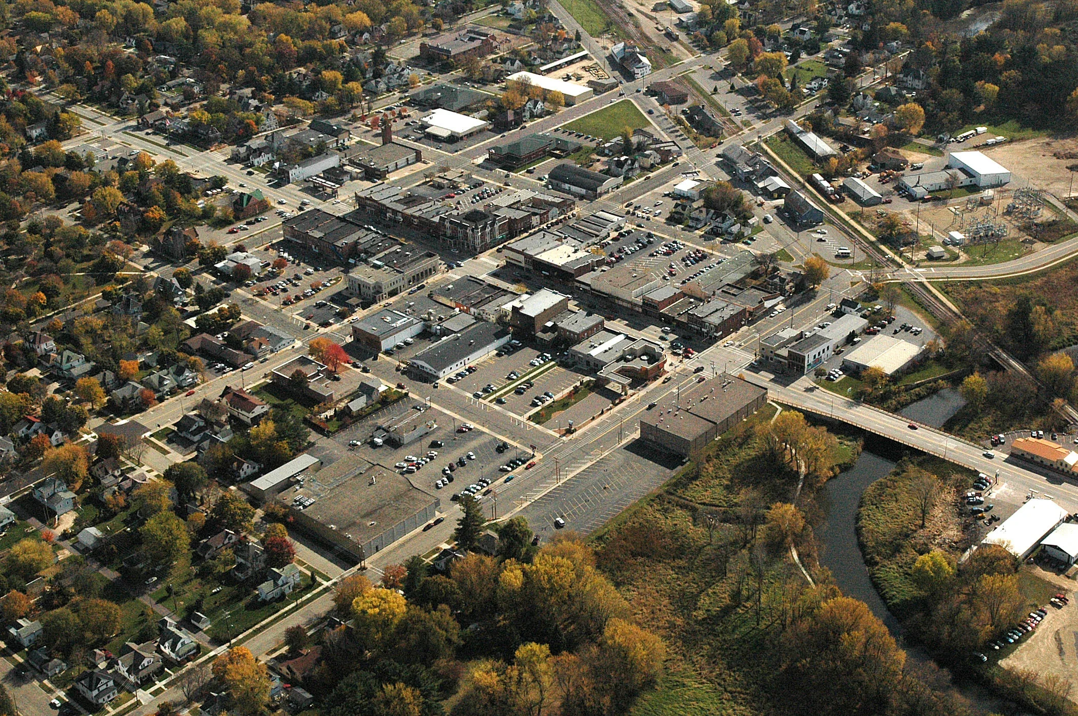 An aerial view of a small town showing a central commercial area with shops and parking lots, surrounded by residential houses, trees with autumn foliage, and a river.