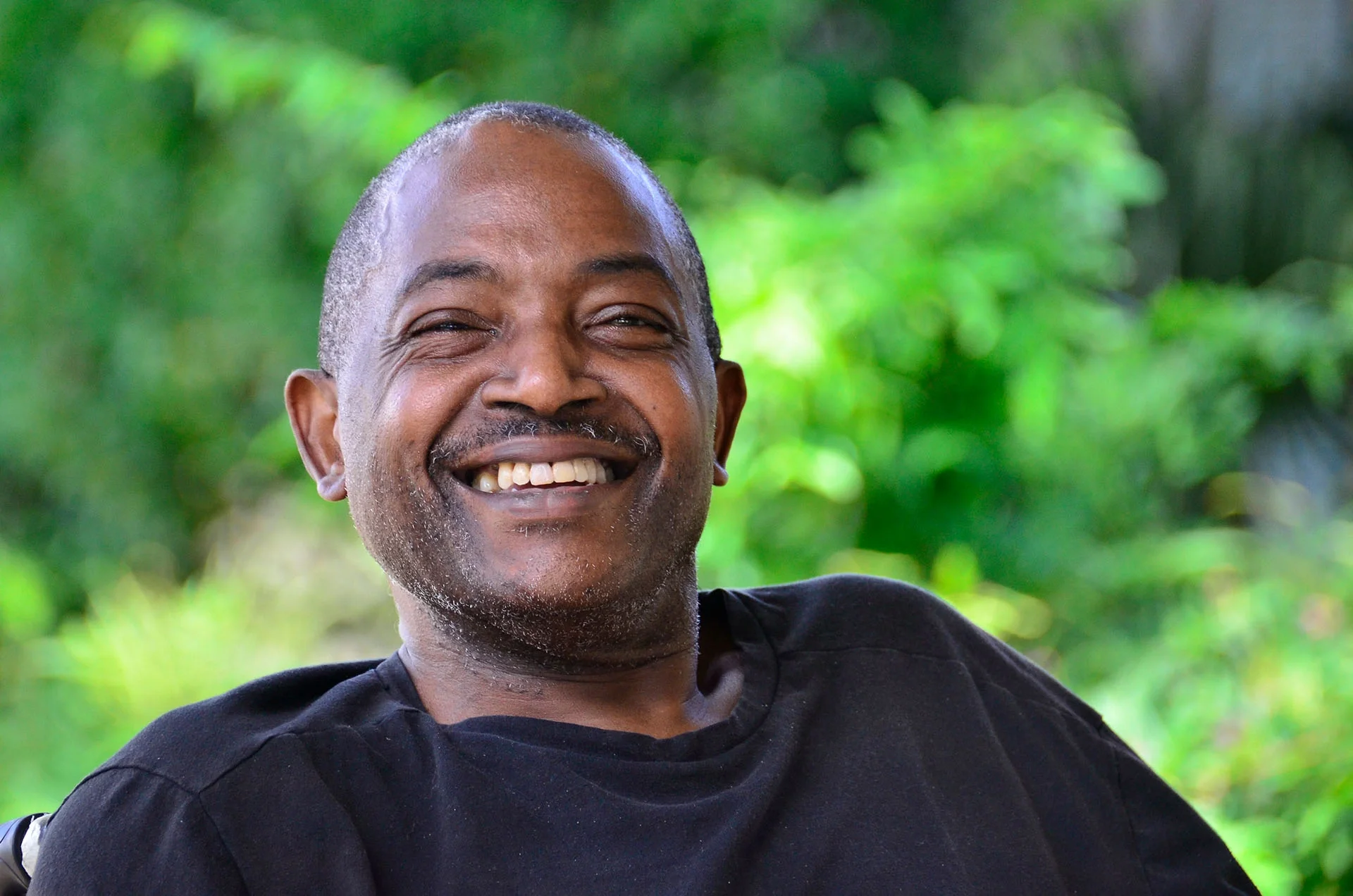 Man smiling outdoors with greenery in background.