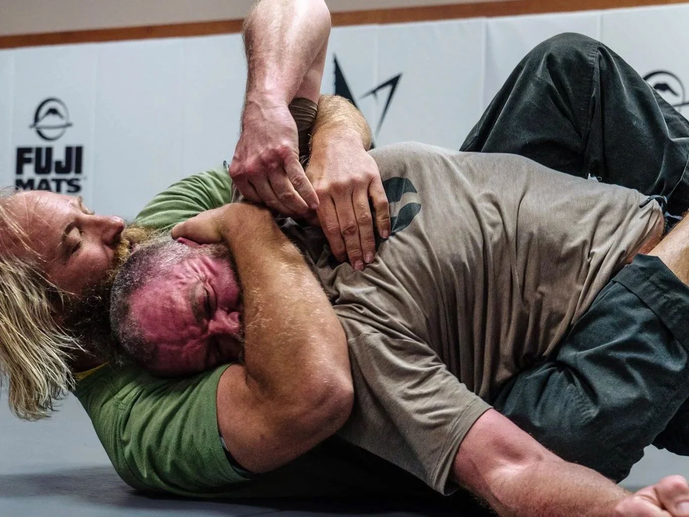 Two men engaged in a wrestling or Brazilian Jiu-Jitsu match on the mat, with one man in a green shirt and the other in a gray shirt, with a gym logo in the background.