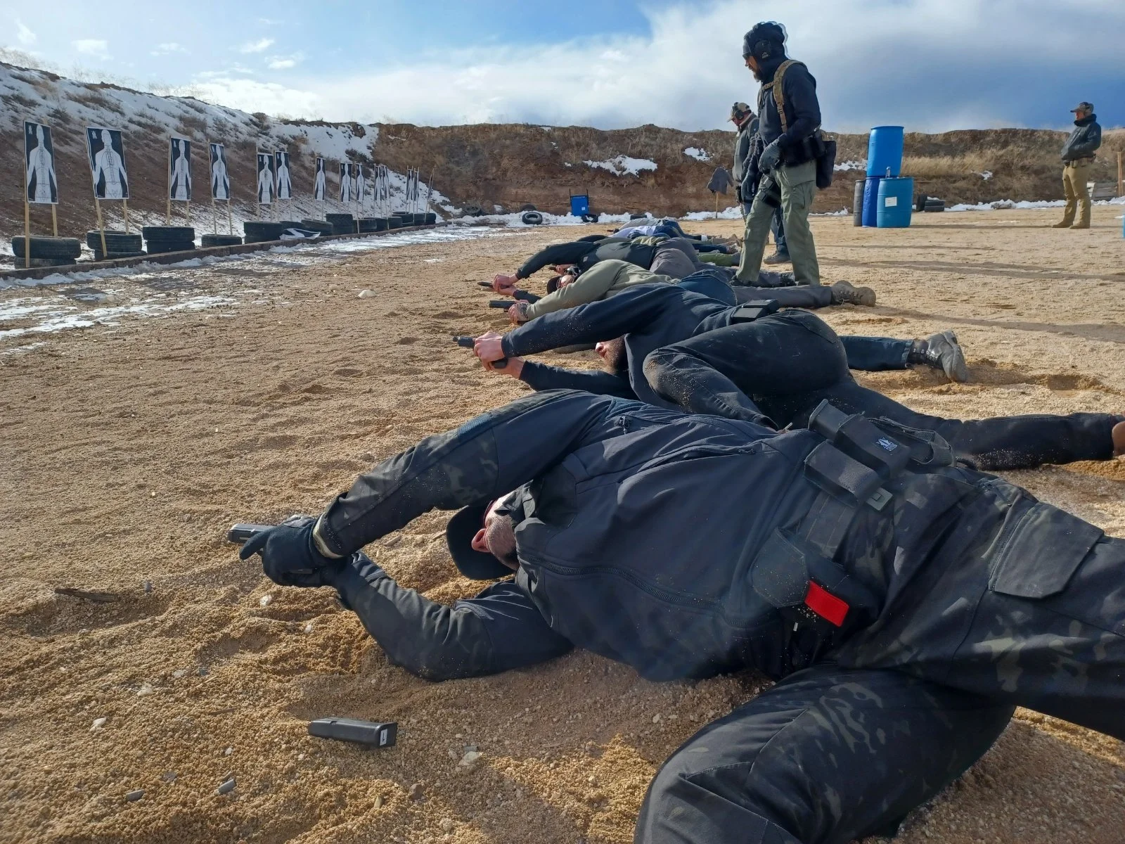 Group of people participating in a firearms training course lying prone on sandy ground, aiming pistols at targets, with several instructors observing, at an outdoor shooting range with snow patches and target stands.