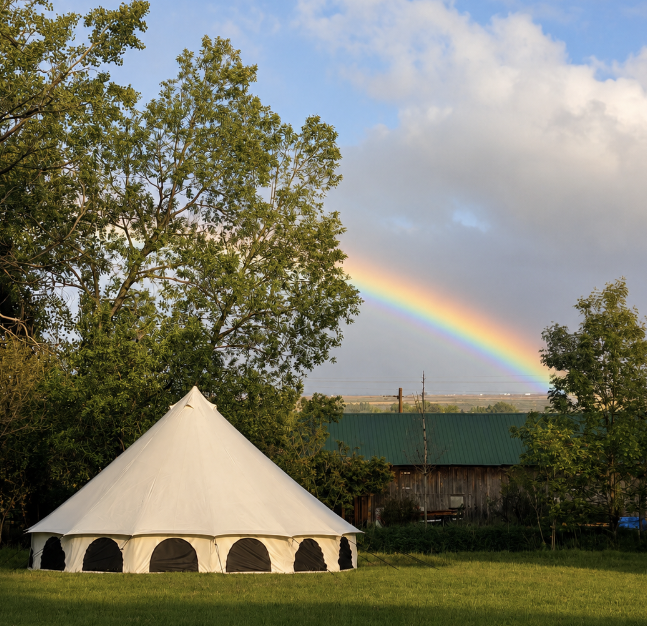 A white tent with black windows set up on a grassy lawn, with trees and a building with a green roof in the background. A rainbow appears in the sky amidst clouds.