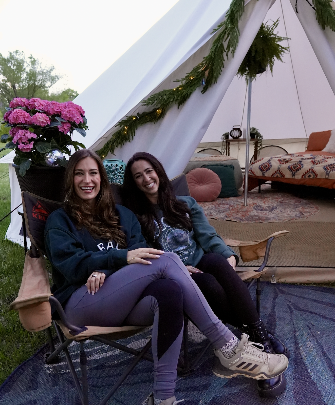 Two women sitting in camping chairs smiling, inside a decorated tent with a Christmas tree and flowers.