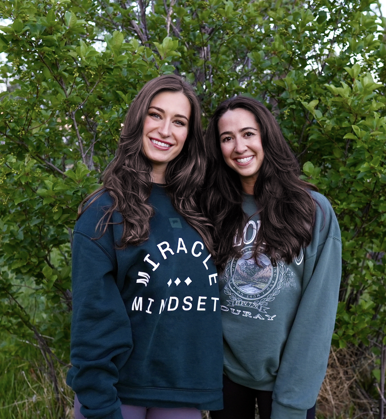 Two women with long dark hair smiling outdoors in front of green leafy bushes.