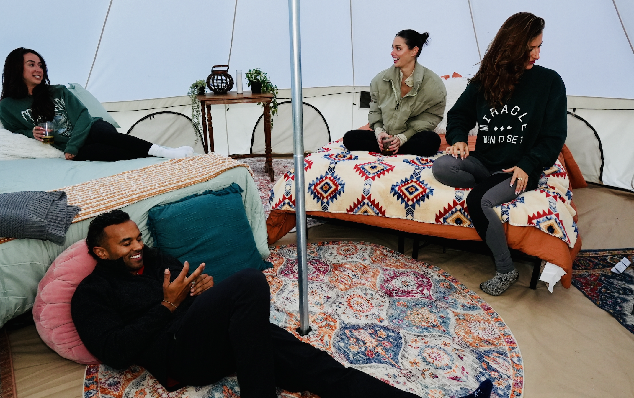Group of people relaxing inside a large tent, sitting on beds and rugs, holding drinks, and engaging in conversation.