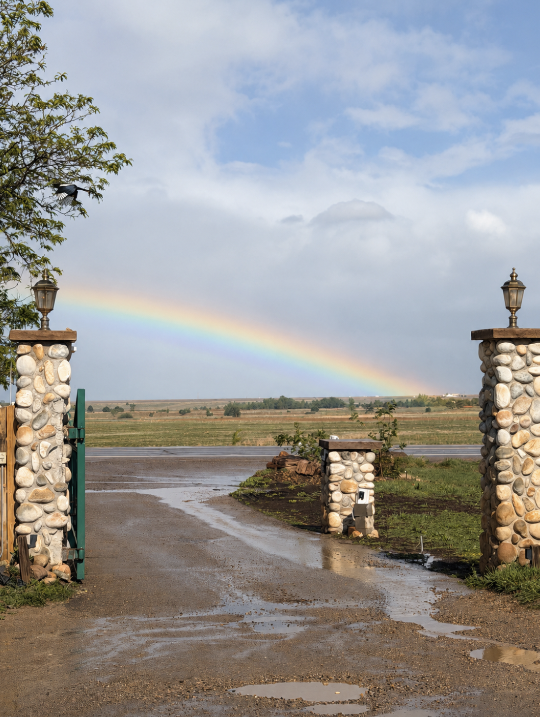 View through an entrance with stone pillars and lamps, showing a muddy driveway leading to a grassy field with a rainbow in the cloudy sky.