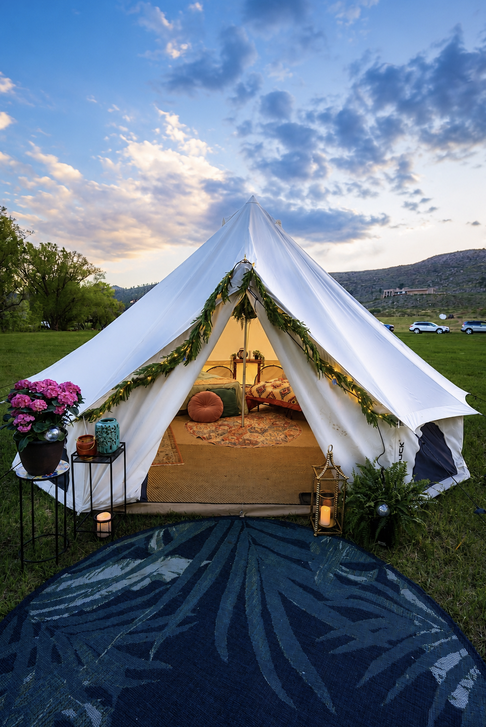 A cozy tent decorated with greenery and fairy lights, with a rug and cushions inside, set up on a grassy field during twilight with a mountain and parked cars in the background.