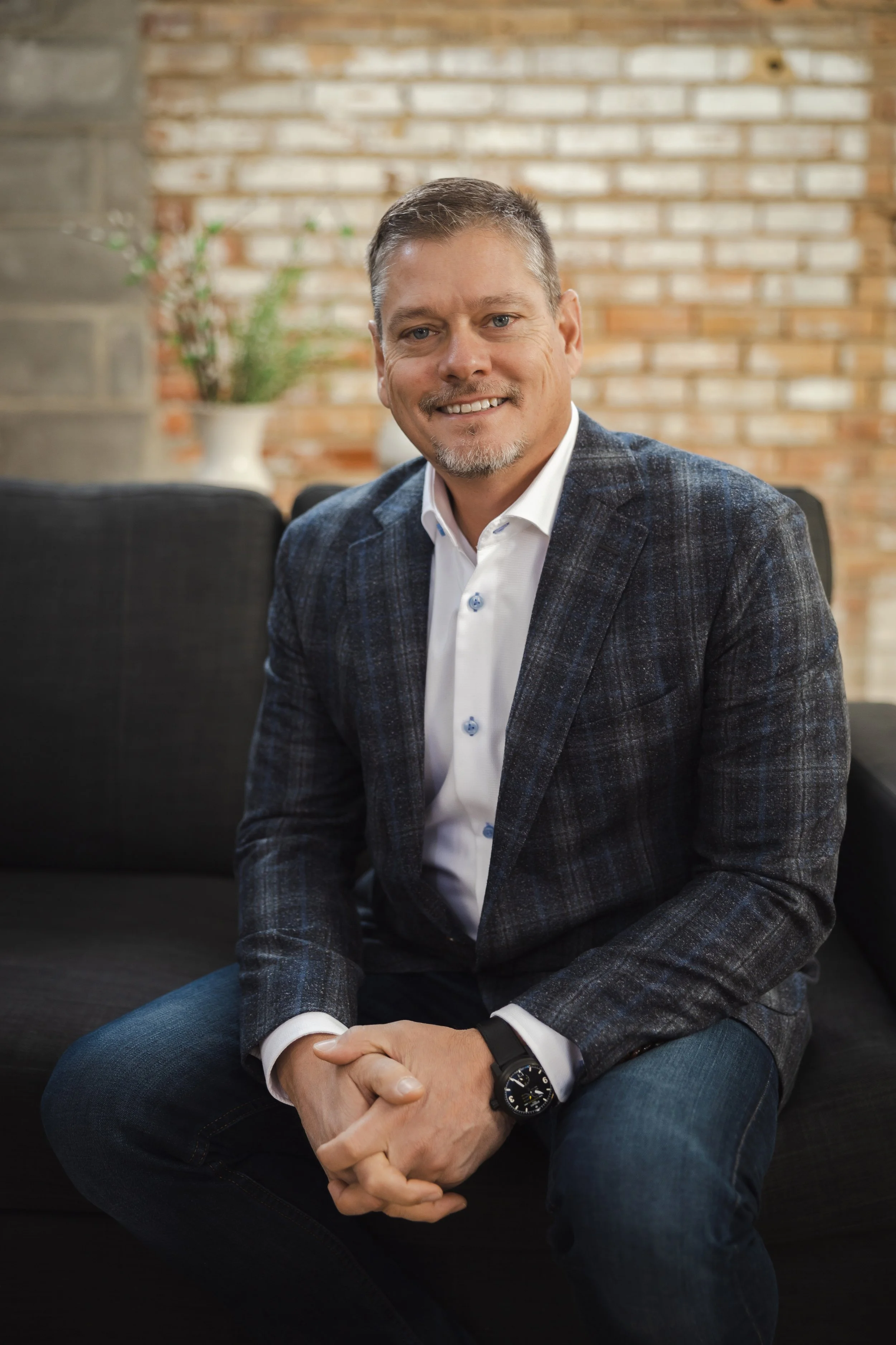 A man with short hair, a beard, and a smile, wearing a dark blazer, white shirt, and jeans, sitting on a black couch in a room with a brick wall background and a white vase with green plants behind him.