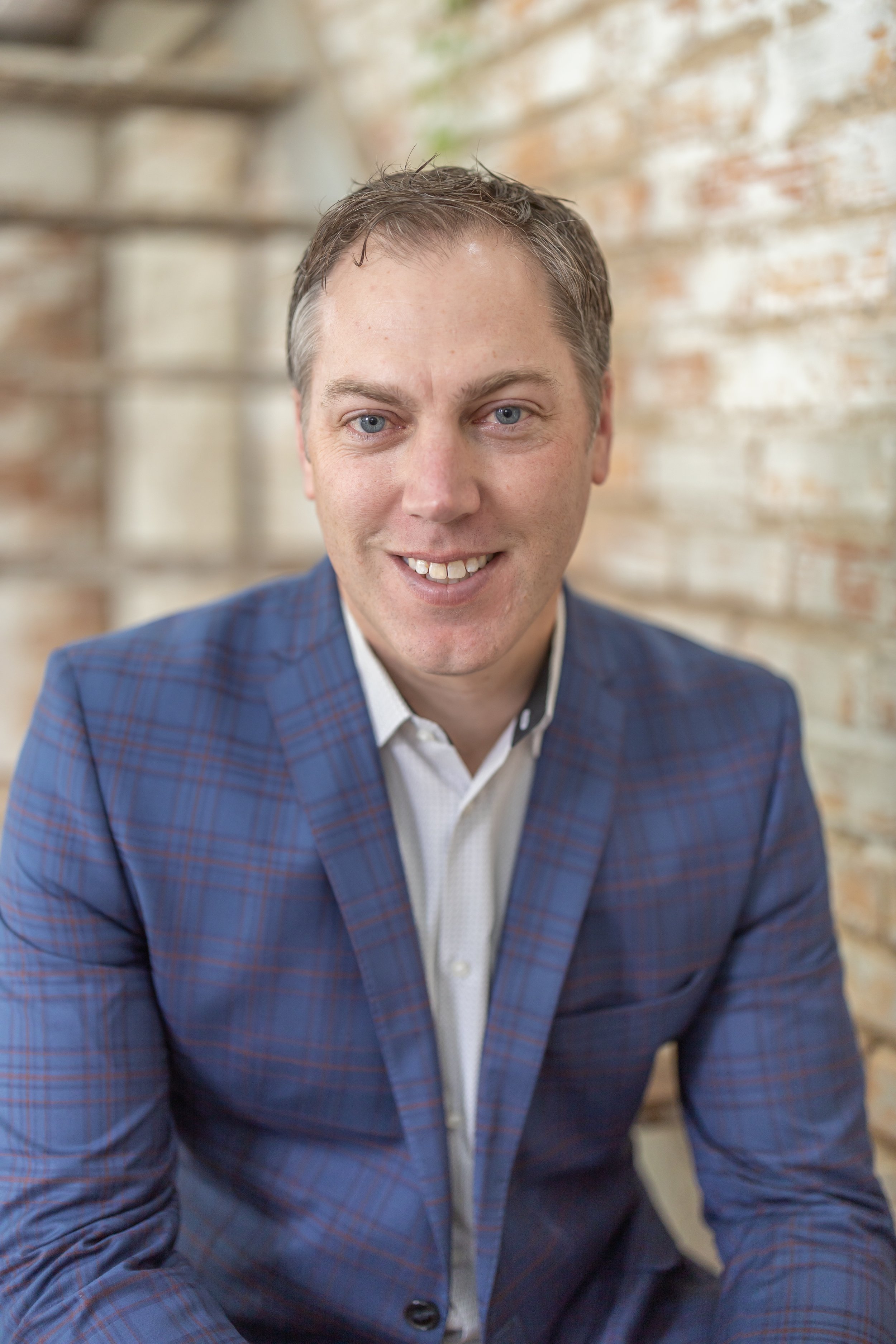 A smiling man with blue eyes and light brown hair, wearing a blue plaid suit jacket and white shirt, sitting in front of a brick wall with blurred wooden shelves in the background.