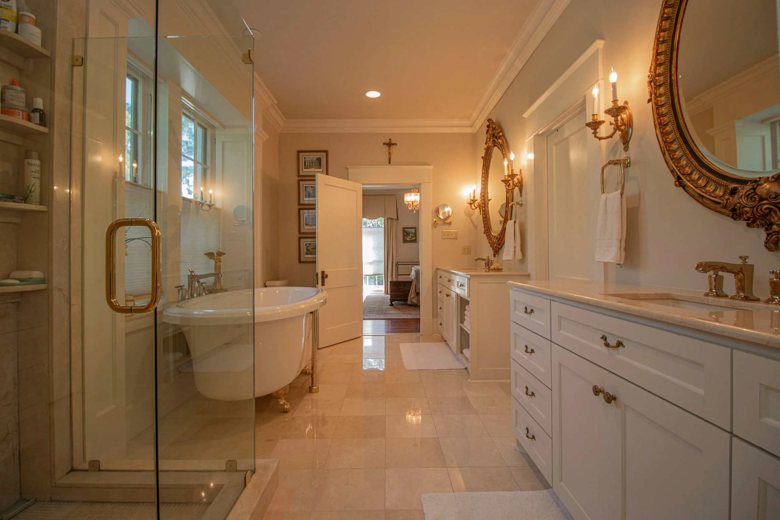 Elegant bathroom with a glass shower, vintage clawfoot bathtub, and white cabinetry with gold fixtures; decorated with framed artwork and ornate mirrors.