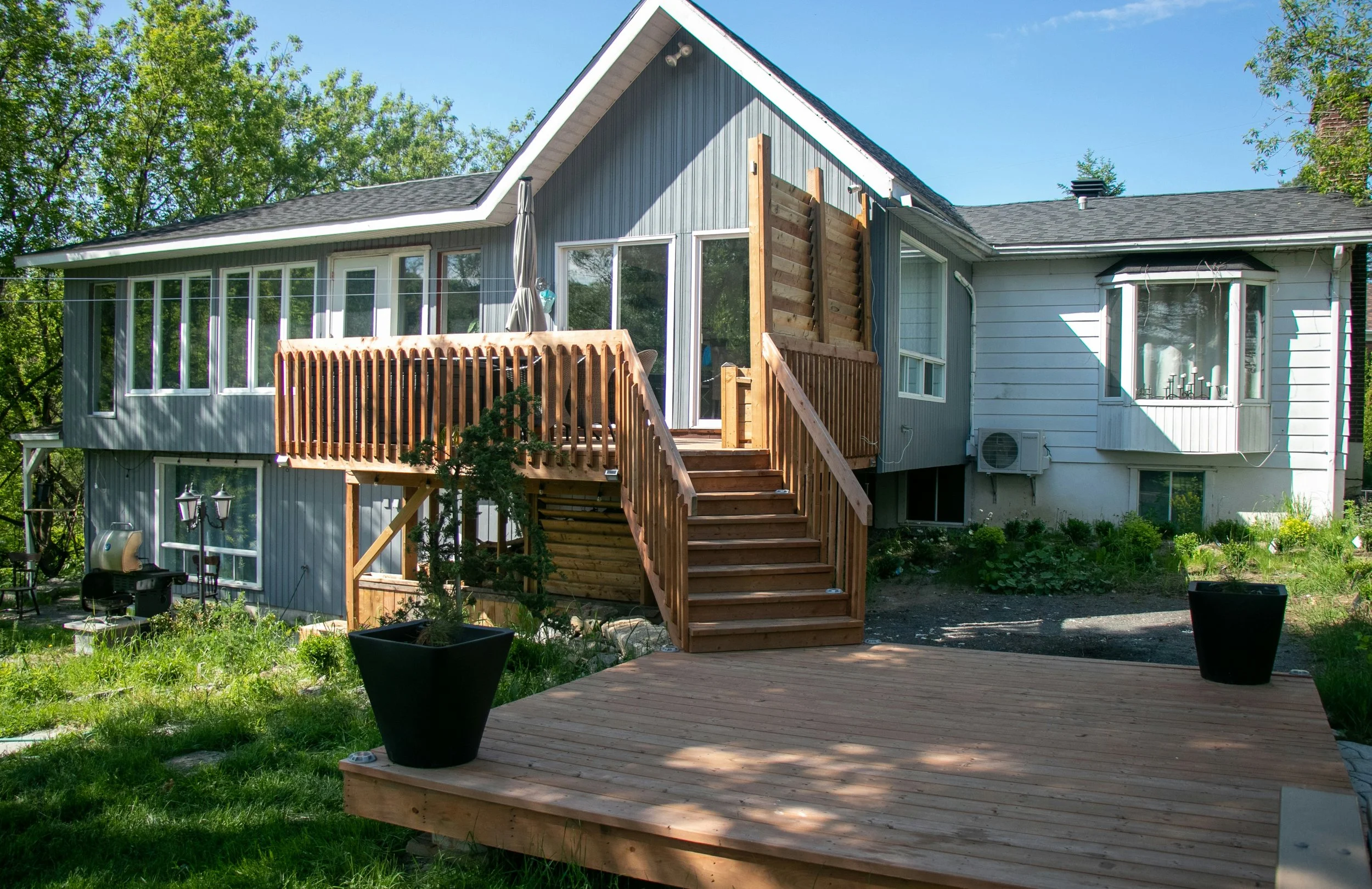 A house with an outdoor wooden deck and stairs leading to a sliding glass door, surrounded by green trees and lawn.