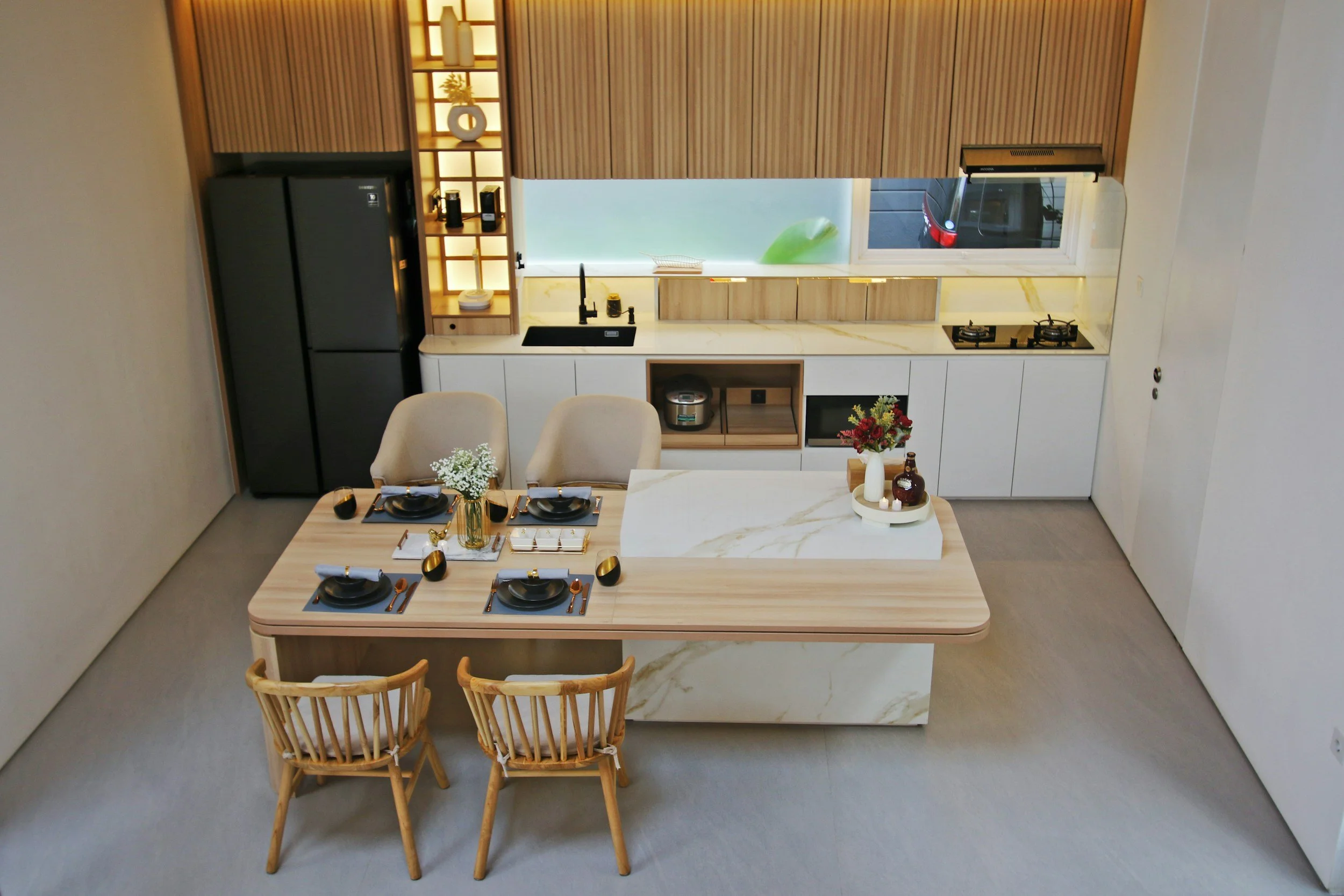 Modern kitchen with a dining area. The kitchen features white cabinets, a black refrigerator, a black sink, a stove, and a window with a view outside. The dining table has four black plates with gold utensils, a vase of white flowers, and a decorativ