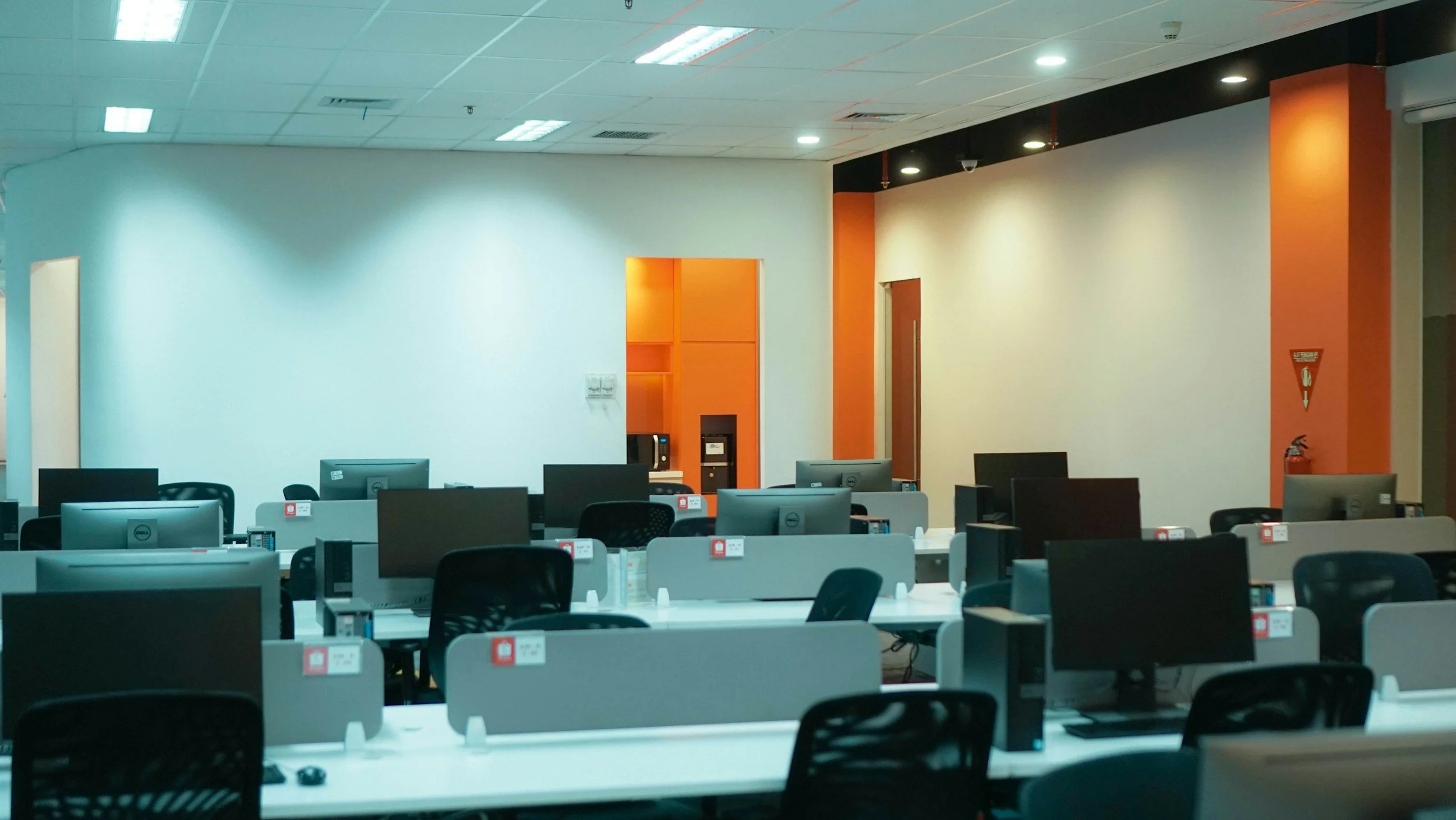 Empty office with rows of desks and computers, orange and white walls, and ceiling lights turned on.
