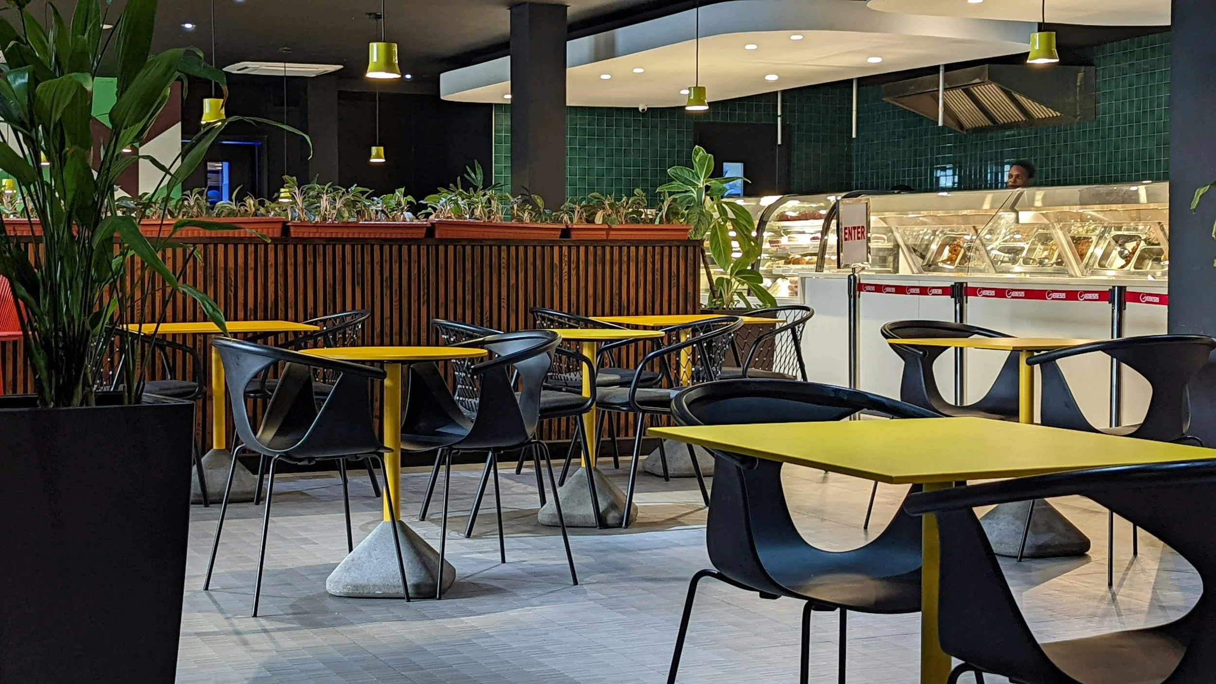 Modern indoor dining area with yellow tables and black chairs, green plants, and a food display counter in the background.