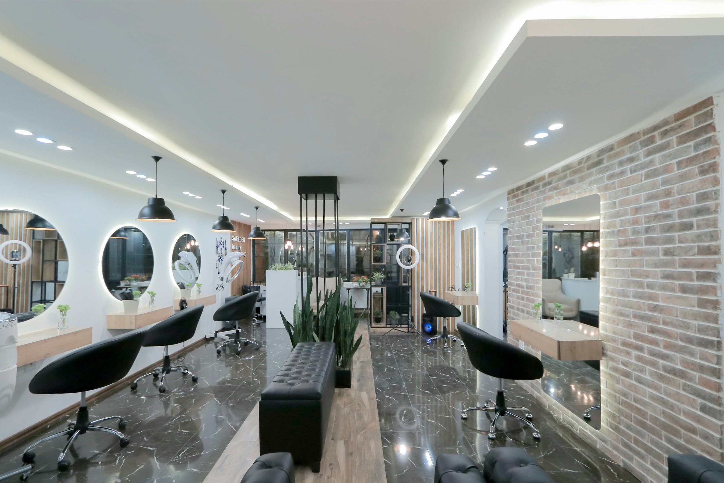 Modern hair salon with black styling chairs, mirrors, and marble flooring. Decor includes round mirrors, hanging pendant lights, and brick wall accents.