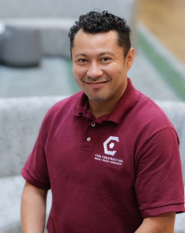 A man with short curly black hair wearing a maroon polo shirt with a logo for Coba Construction, smiling and sitting outdoors.