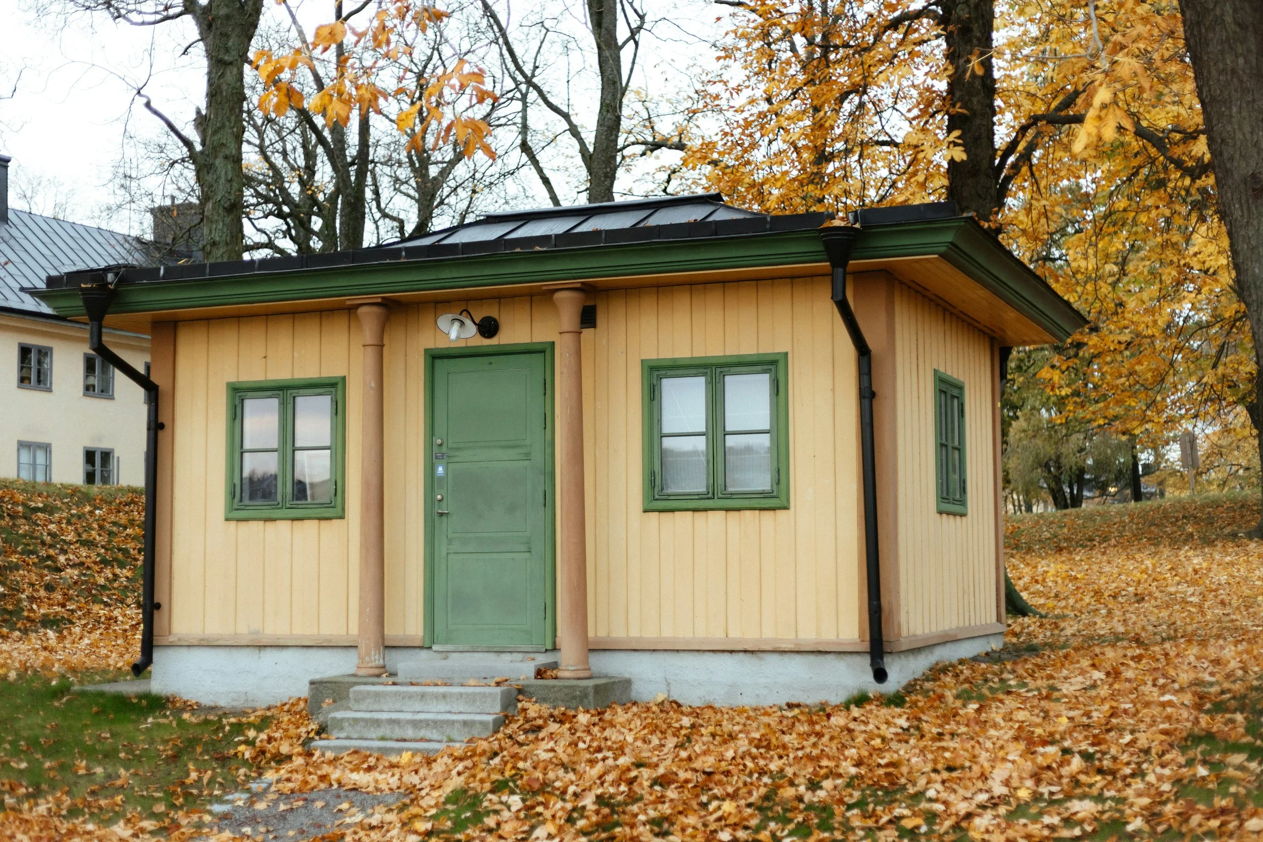 Small yellow wooden house with green trim, green door, two windows, and a small set of stairs, surrounded by autumn leaves and trees.