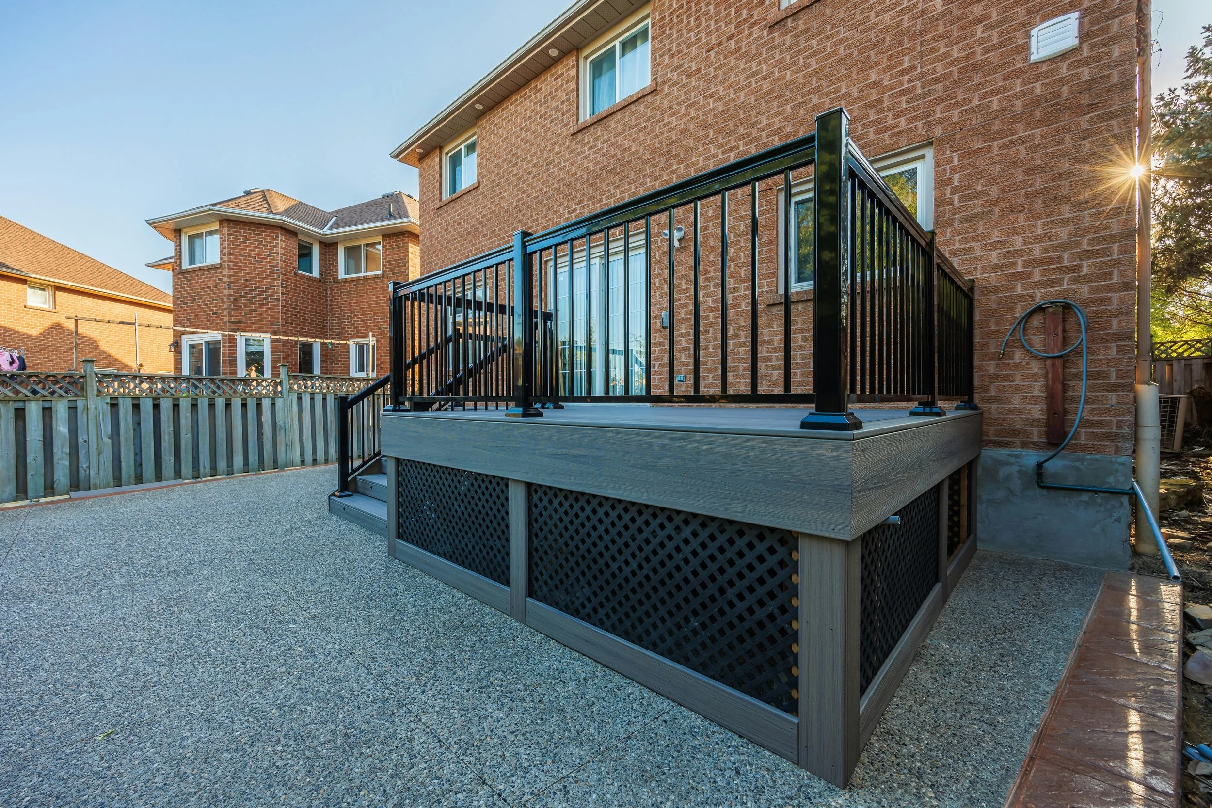Backyard patio with newly built wooden deck and black metal railing attached to a brick house. The patio is surrounded by a wooden fence, and other two-story brick houses are visible in the background. The sun is setting behind the trees, casting a w