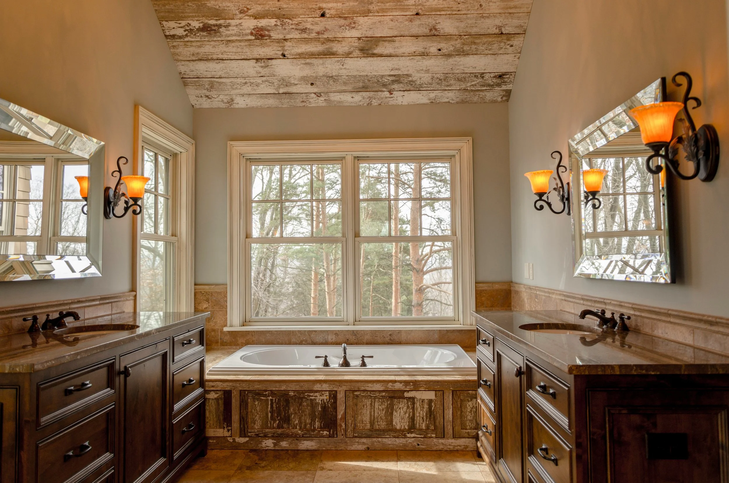 A spacious bathroom with double dark wood vanities, each with a sink, located on either side of a large window. There is a bathtub beneath the window, and the ceiling is made of rustic, weathered wood. Two wall-mounted mirrors and four wall sconces w
