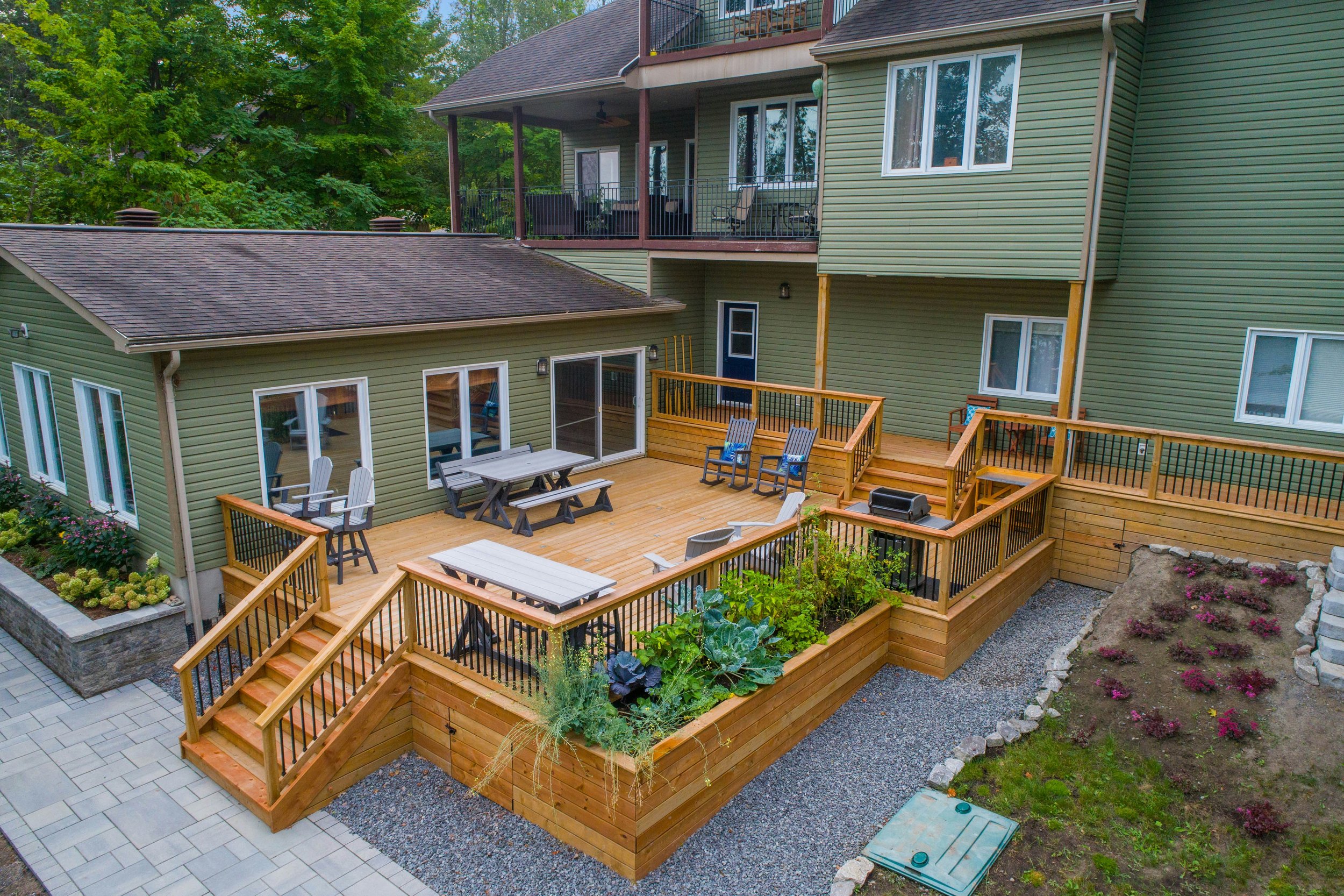 View of a multi-level backyard deck attached to a green house, with outdoor furniture, potted plants, and an area with flowering plants and gravel. The house has large windows and sliding glass doors, and there's a balcony on the upper floor.