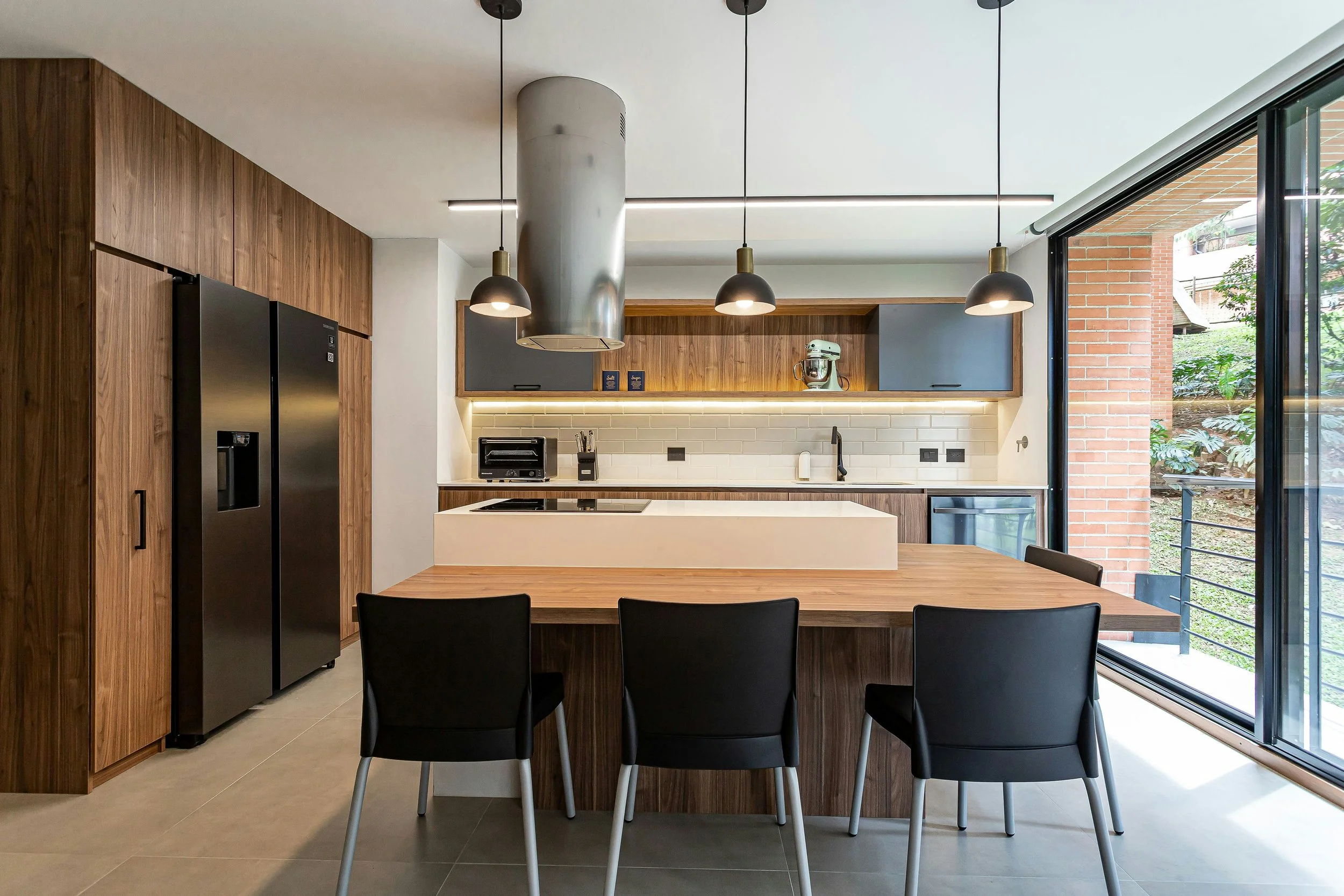 Modern kitchen with wooden cabinets, black appliances, and a dining table with four chairs, large sliding glass door opening to a balcony, pendant lighting, and a white brick backsplash.
