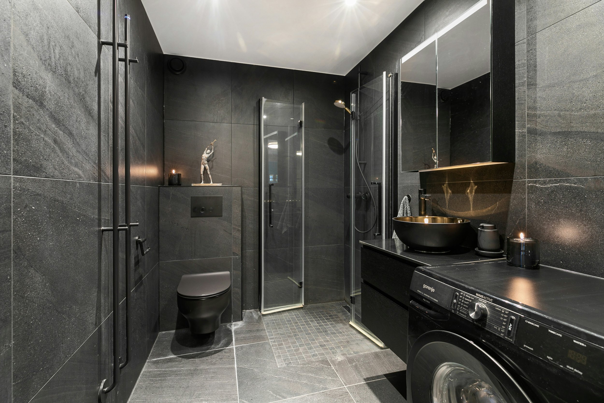 Modern black-tiled bathroom with a toilet, shower, and black sink with gold interior, a mirror cabinet, and a washing machine.