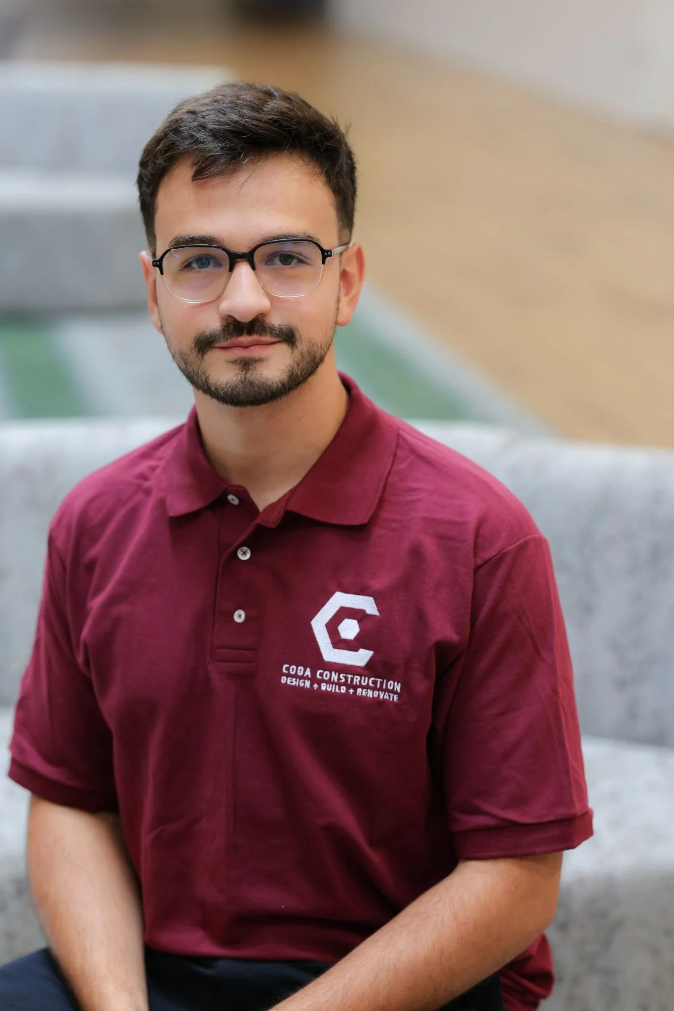 A young man with dark hair, glasses, and a beard is sitting on a grey couch in an indoor setting. He is wearing a maroon polo shirt with a logo and text that reads 'Coba Construction' and 'Design + Build + Renovate'.
