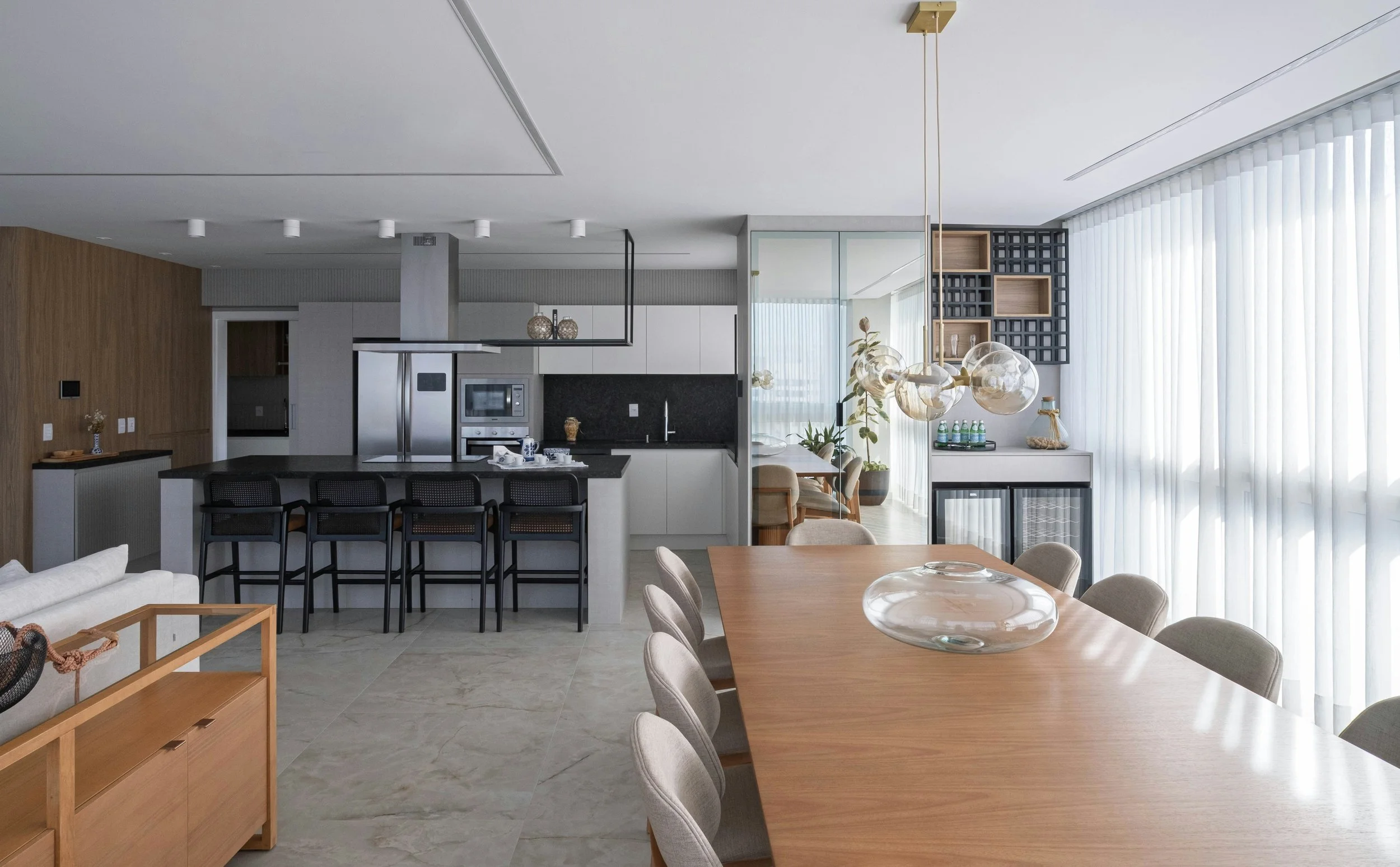 Modern open-concept living space featuring a kitchen with a black countertop island, right of a dining area with a large wooden table and beige chairs, and a cozy breakfast nook with seating by large windows with sheer curtains. Decor includes natura