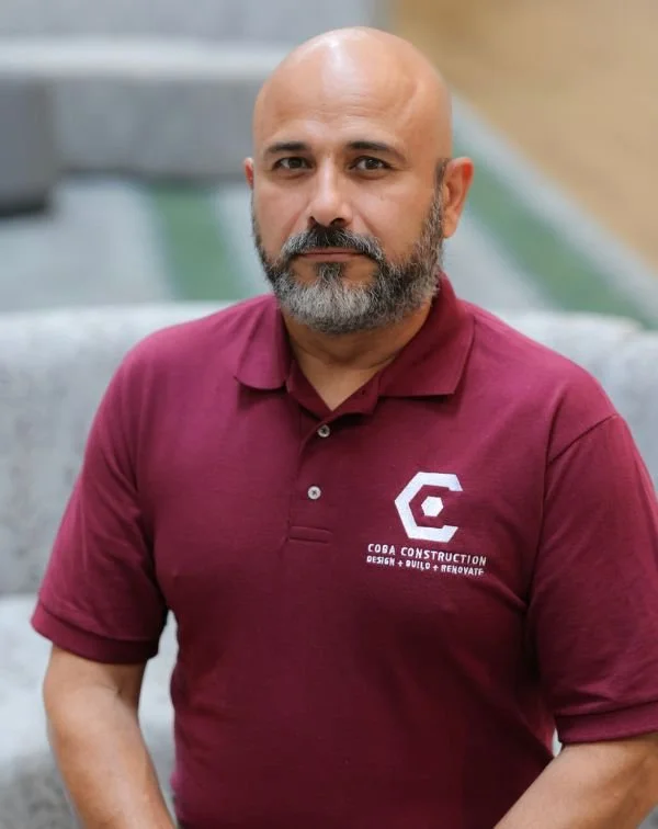 A man with a shaved head and beard wearing a maroon polo shirt with the logo and text 'Coda Construction, Design, Build, Renovate' standing in front of a modern building with concrete and glass features.