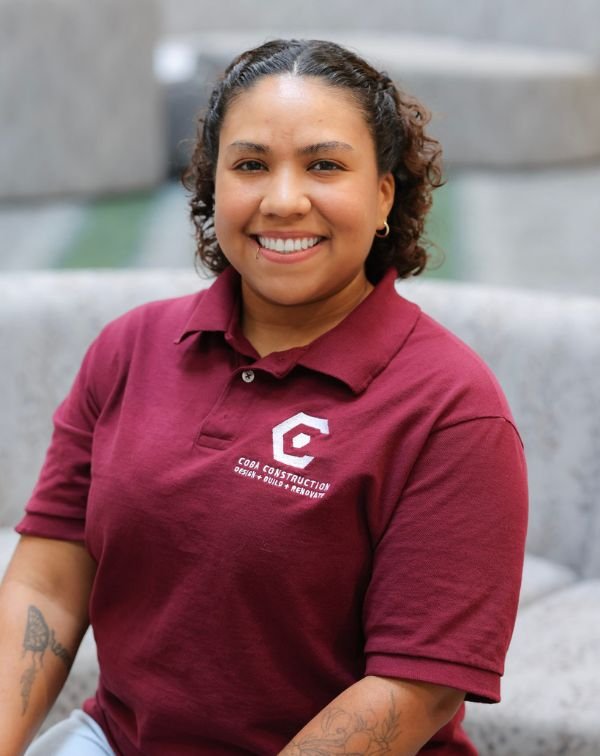 A woman with curly hair smiling, wearing a maroon polo shirt with a logo and text, sitting on gray outdoor seating.