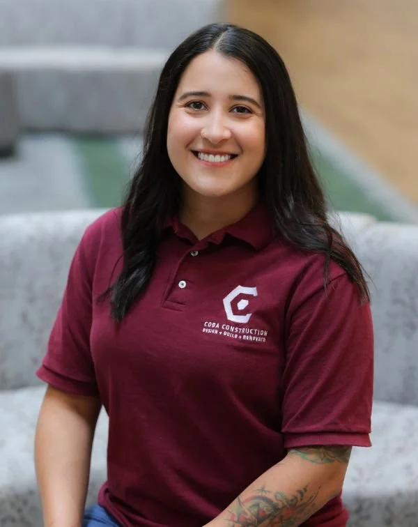 A woman with long dark hair, smiling, wearing a maroon polo shirt with a logo and text that reads 'Coda Construction Design Build Renovate,' sitting on a gray couch in a modern indoor setting.