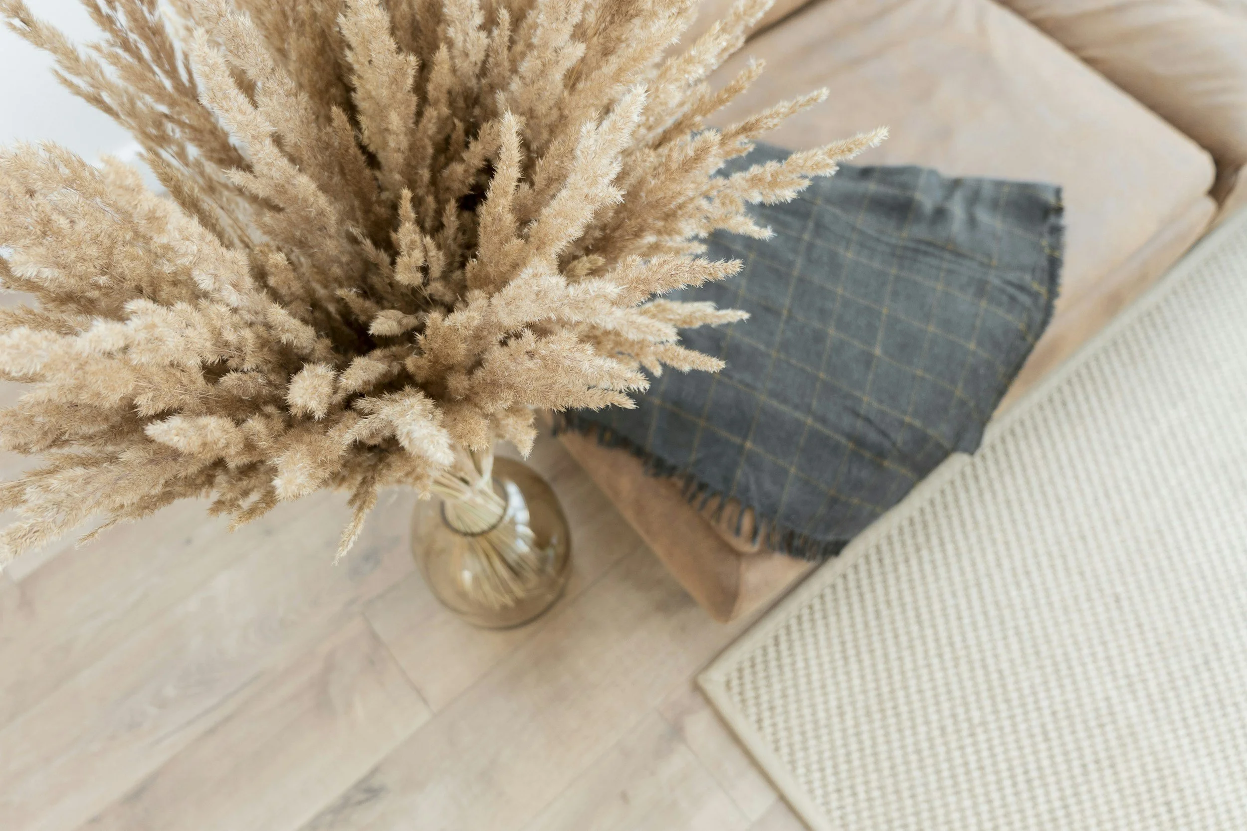 A top-down view of a living room corner with a beige sofa, a gray plaid blanket, a beige cushion, and a glass vase with dried pampas grass on a wooden floor.