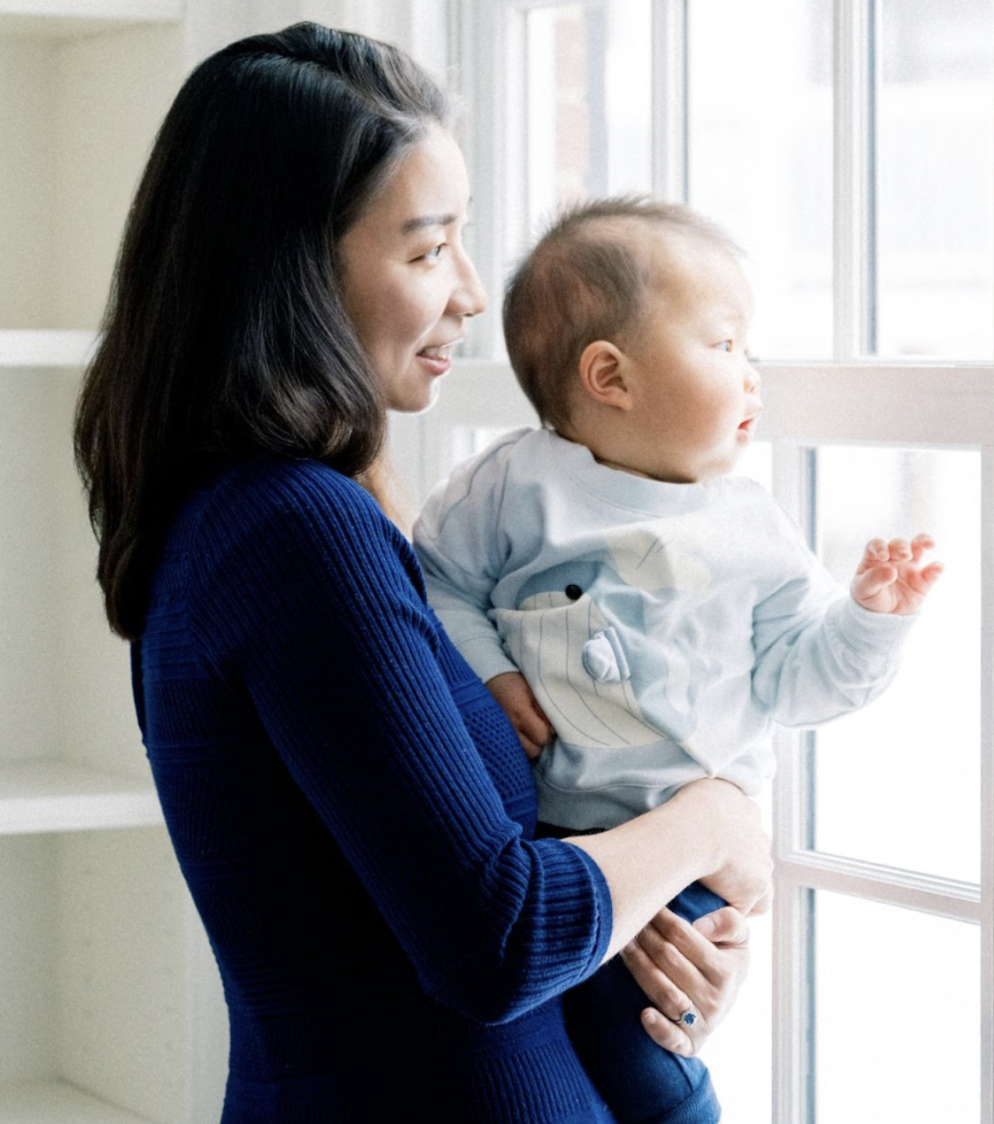 A woman with dark hair wearing a blue sweater holds a young child with light hair wearing a white shirt as they look out a window together.