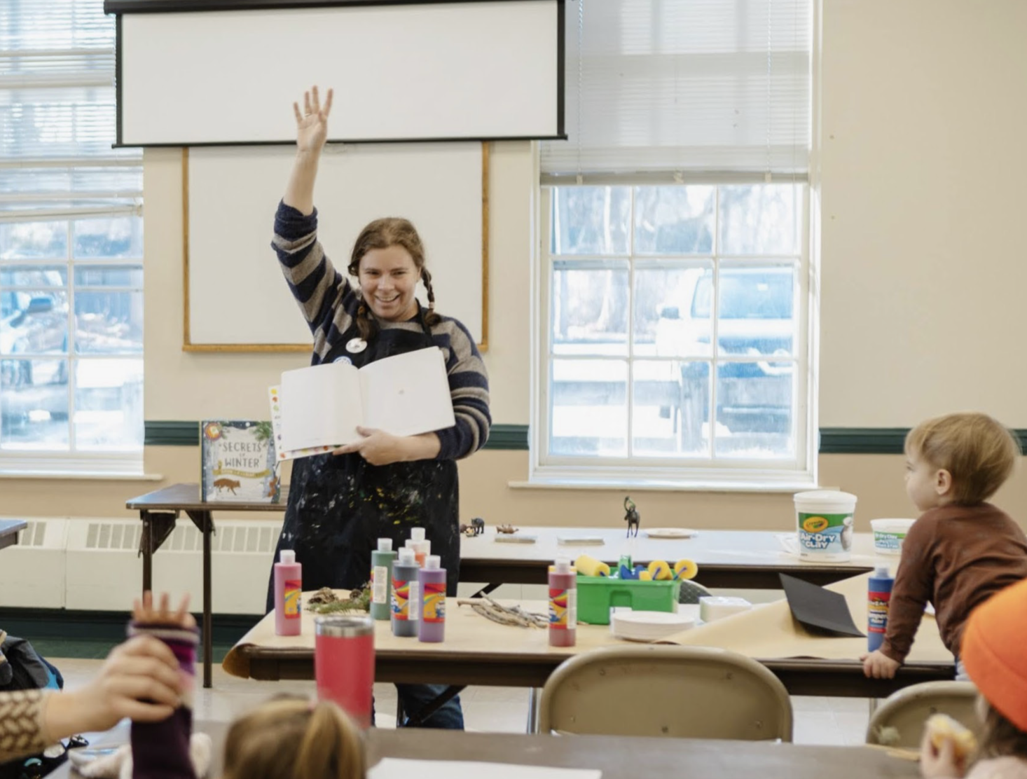 A woman standing in front of a classroom holding an open book and smiling, with her right hand raised. Children are seated at tables in front, with art supplies and small figurines on the tables. Large windows in the background with a snowy landscape outside.