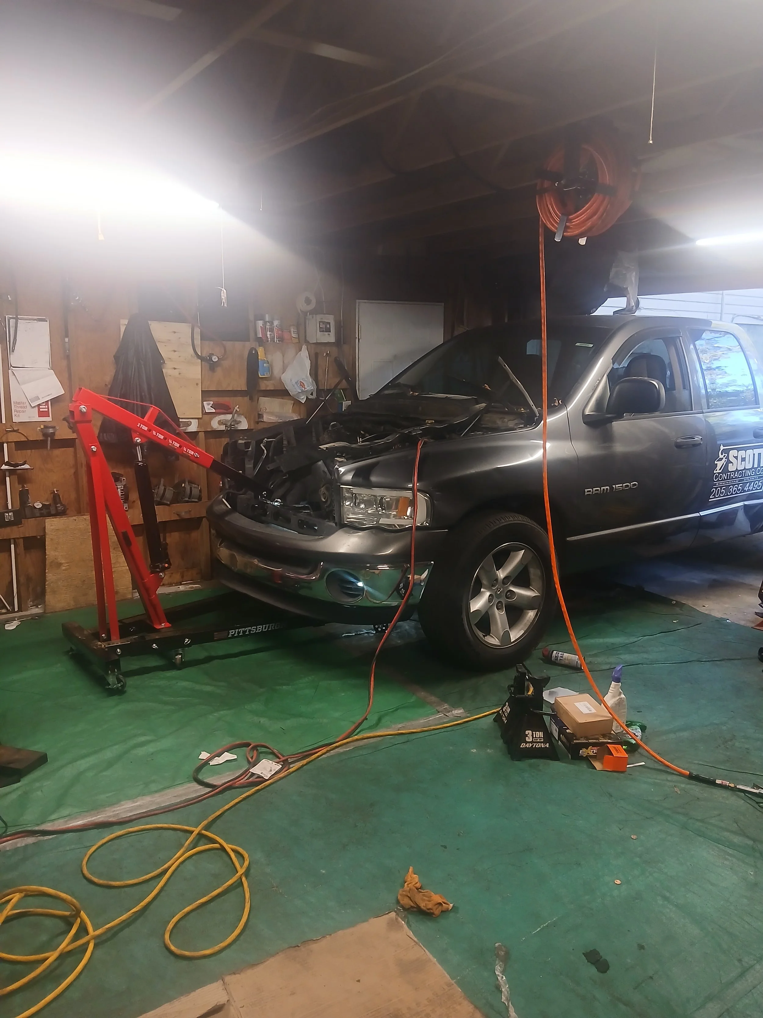 A silver Dodge Ram 1500 truck inside a garage being repaired, with the front hood open and a red engine hoist in front of it. Various tools and items are hanging on the wooden wall and on the floor.