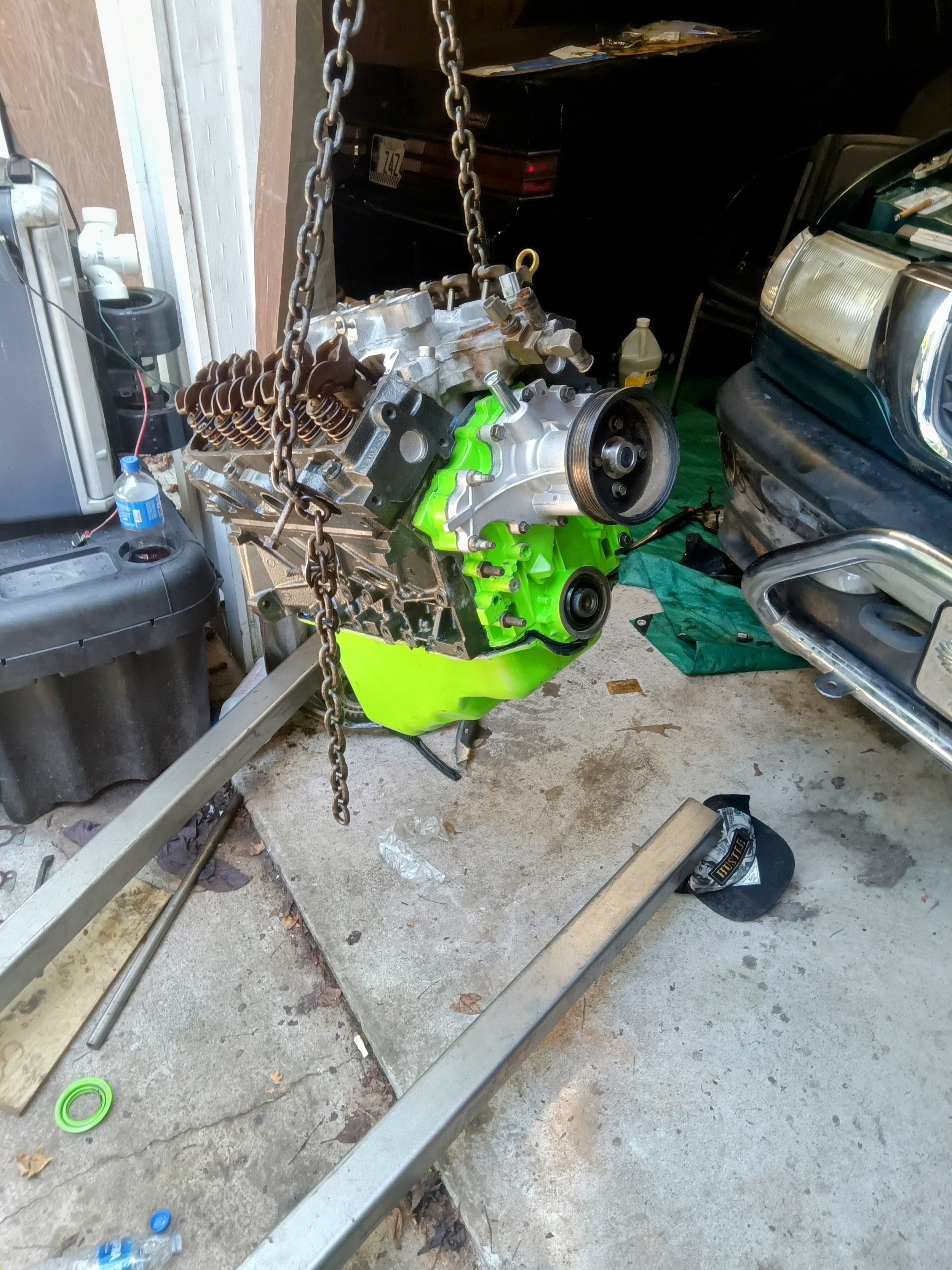 A car engine is suspended by a chain hoist in a garage, surrounded by various tools and car parts, with a vehicle parked nearby and some bottles and containers on the ground.