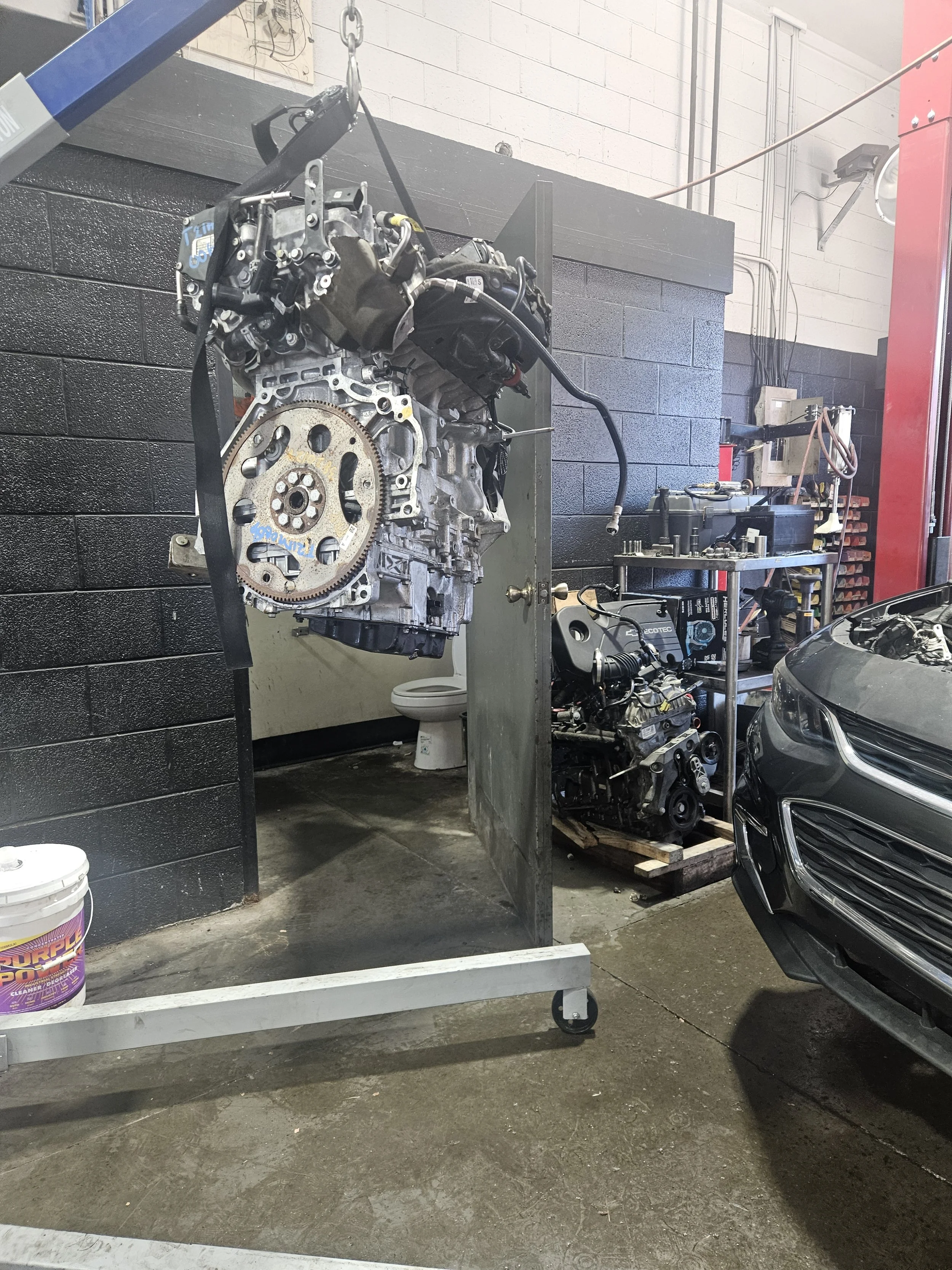 An automotive workshop with a car engine hoisted and hanging in the air, a car partially visible on the right, and various tools and equipment on shelves in the background.