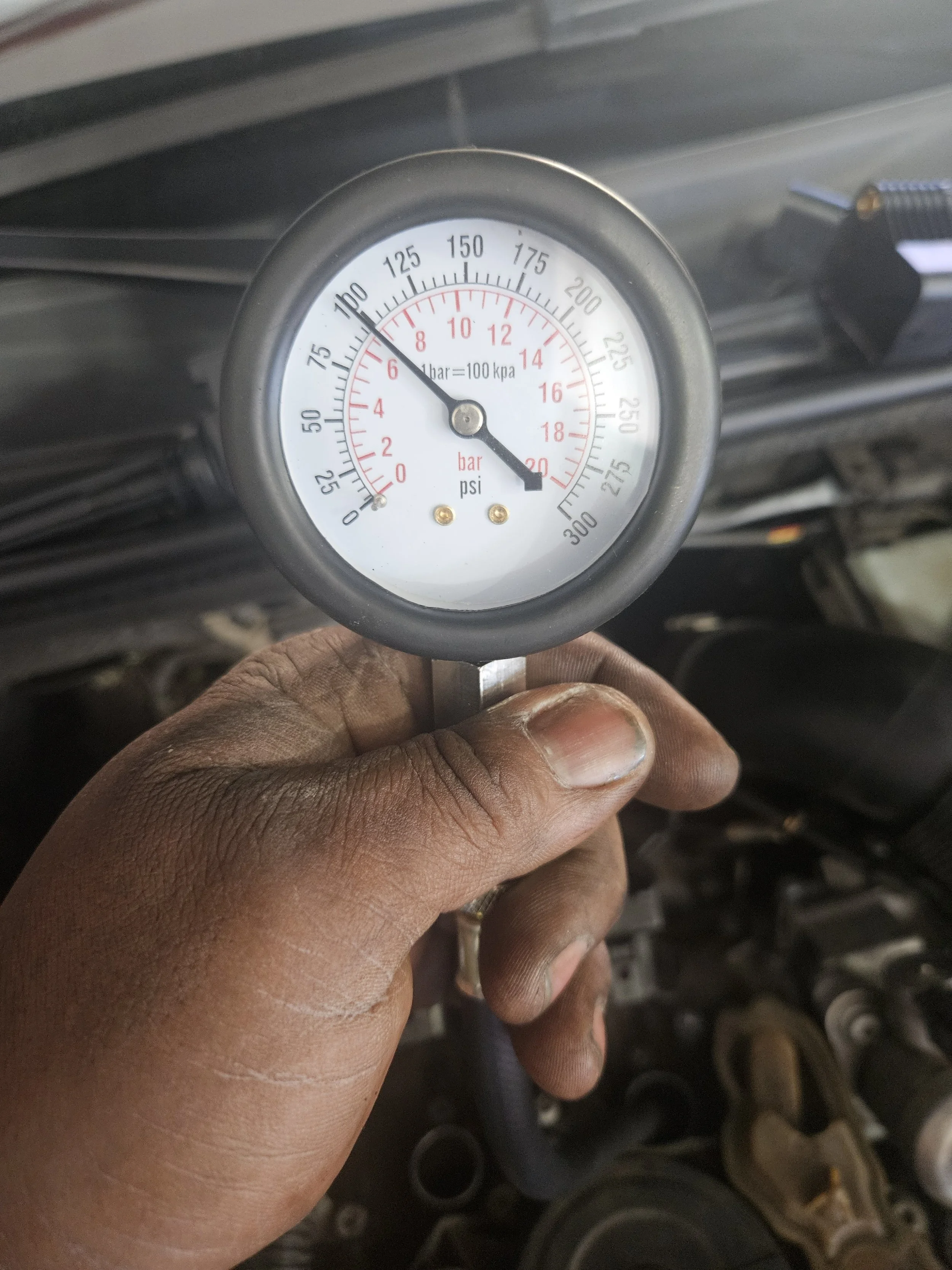 Hand holding a pressure gauge showing approximately 8 bar or 120 psi in a car engine bay.